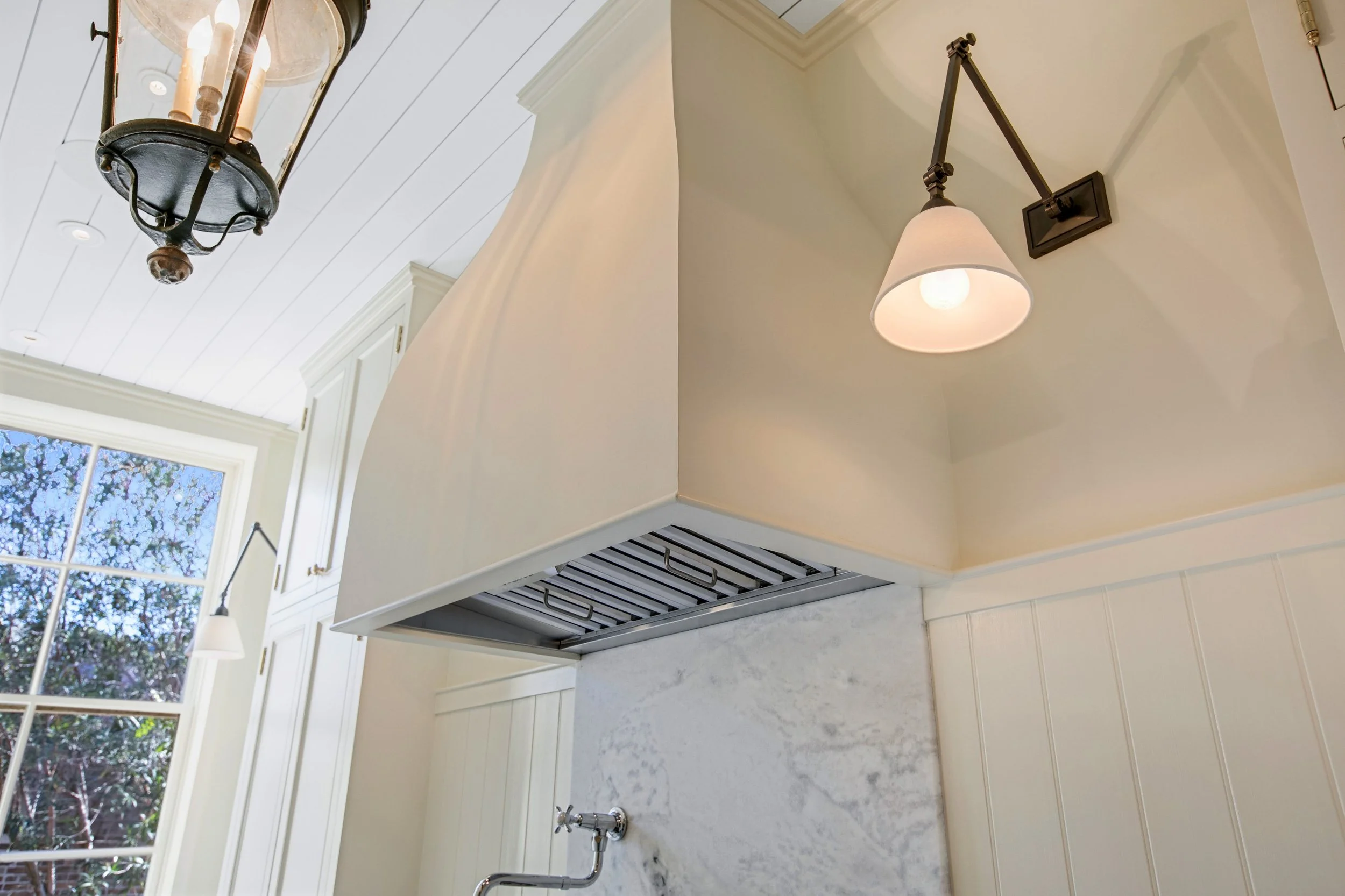 Interior view of a kitchen ceiling showing a chandelier, a wall-mounted light fixture, and a vent hood over a marble backsplash near a window with trees outside.