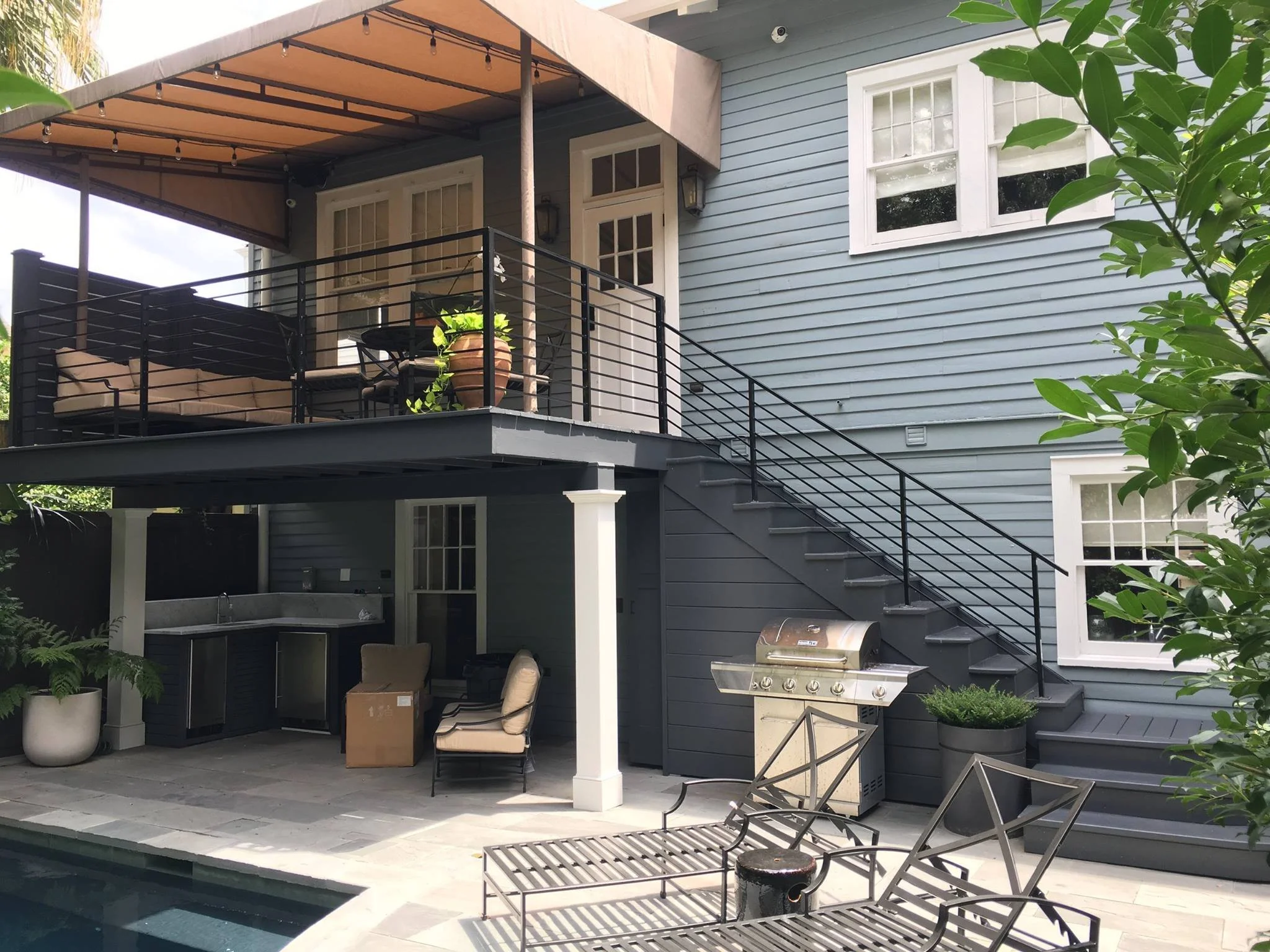 A backyard patio and balcony area at a modern house, featuring outdoor furniture, a countertop with a sink, a gas grill, potted plants, and stairs leading to the upper deck. The house has blue siding and white-framed windows.