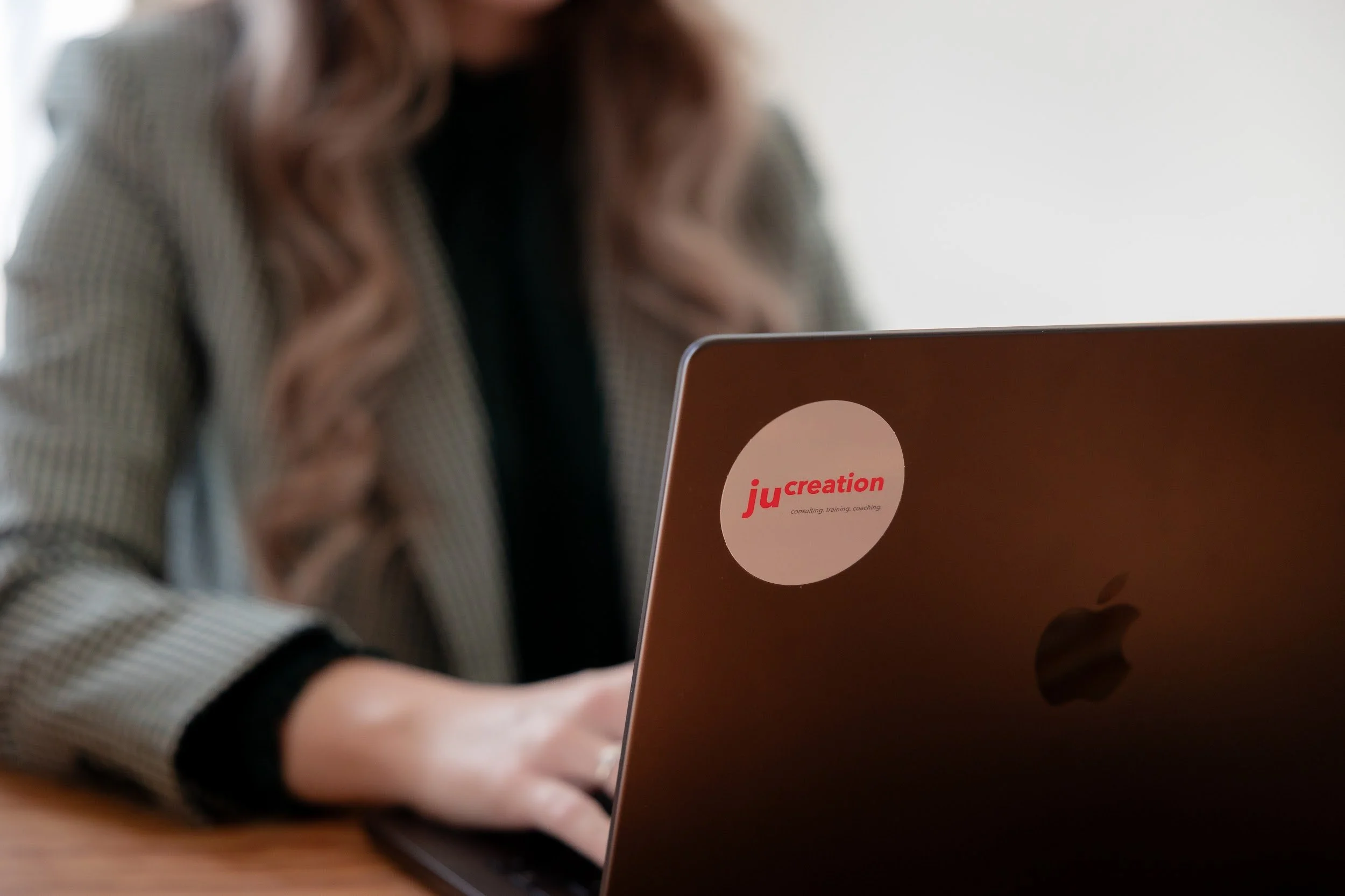 Leonie Gröschner sitzt an einem Tisch, die Kamera fokussiert auf den oberen Teil eines Laptops mit einem Aufkleber, auf dem ‚jucreation‘ steht, während sie an einem Schreibtisch arbeitet.
