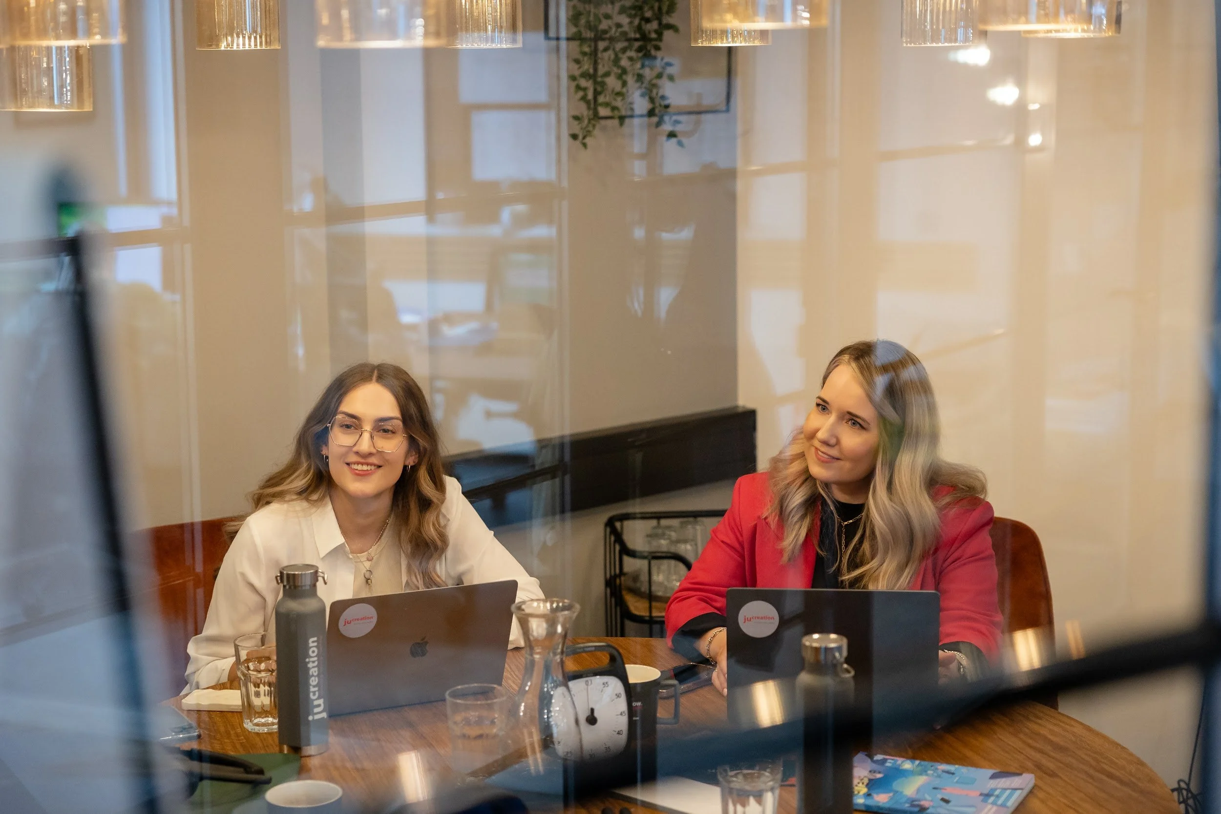 Juliane Superka und Leonie Gröschner sitzen an einem Tisch in einem Besprechungsraum, Blick durch eine Glasscheibe.