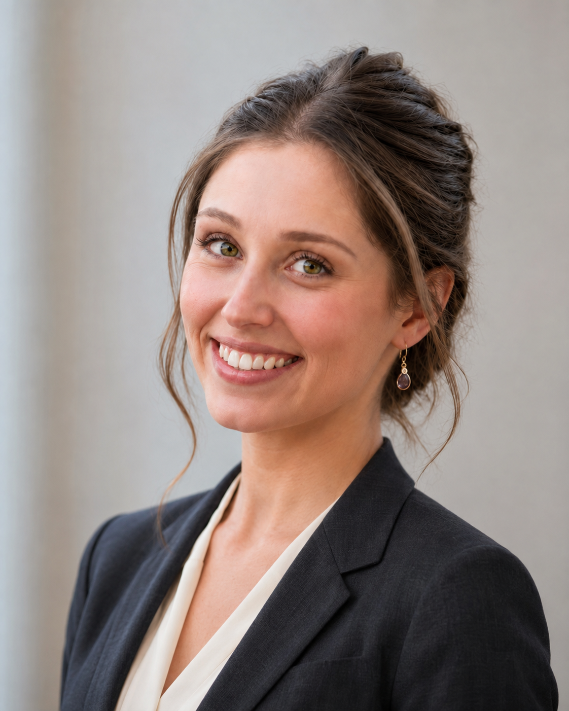Professional woman smiling, wearing a dark blazer and earrings, with light makeup and styled hair, against a neutral background.
