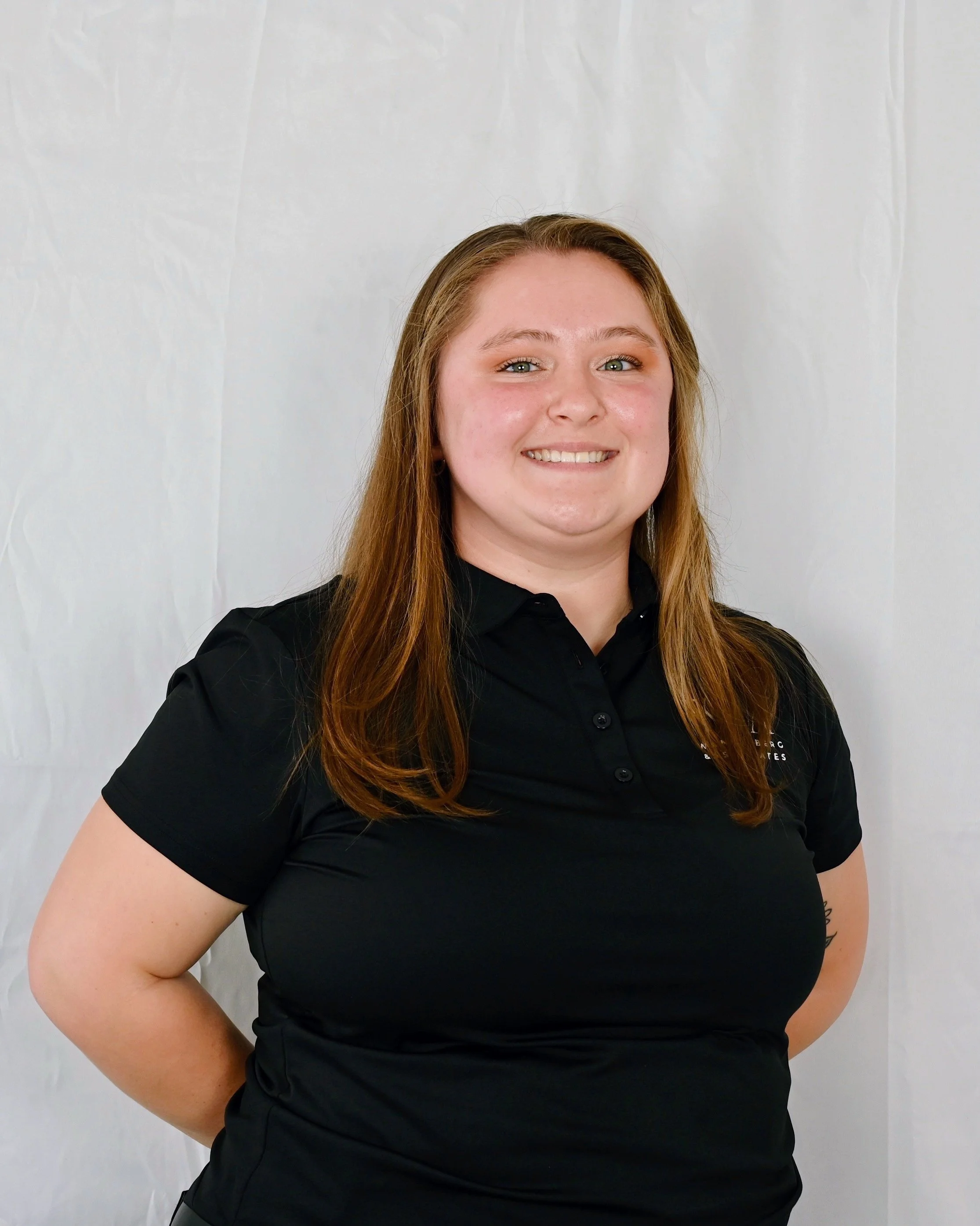 A woman with long red hair wearing a black polo shirt, smiling and standing against a plain white background.