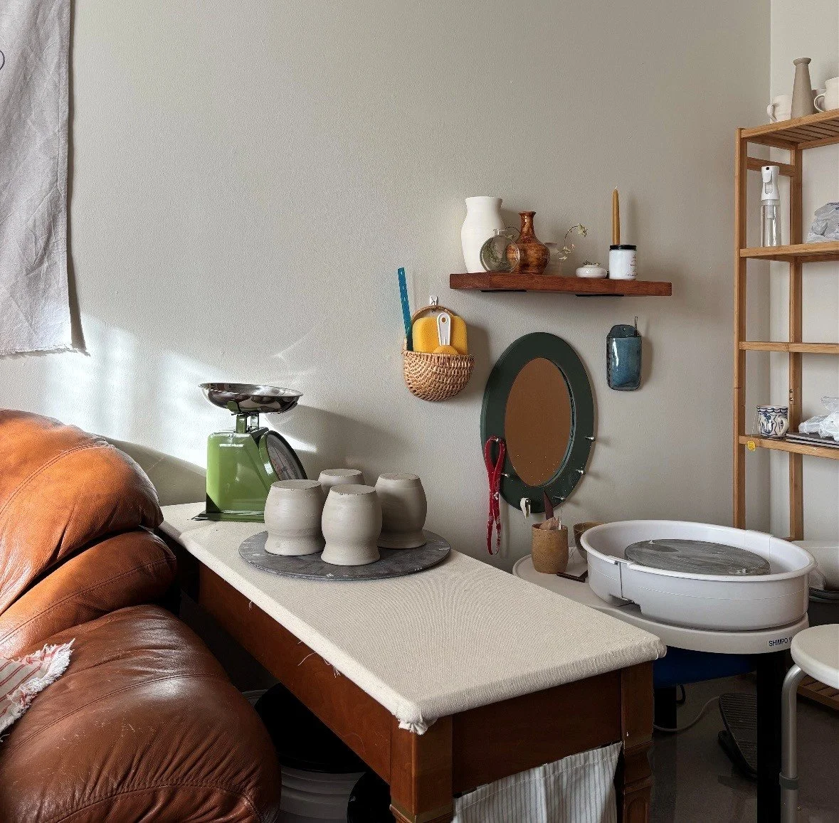 A room with pottery, a vintage scale, a mirror, and a clay sculpture, with shelves holding vases and a plant, cozy lighting, and a leather sofa.