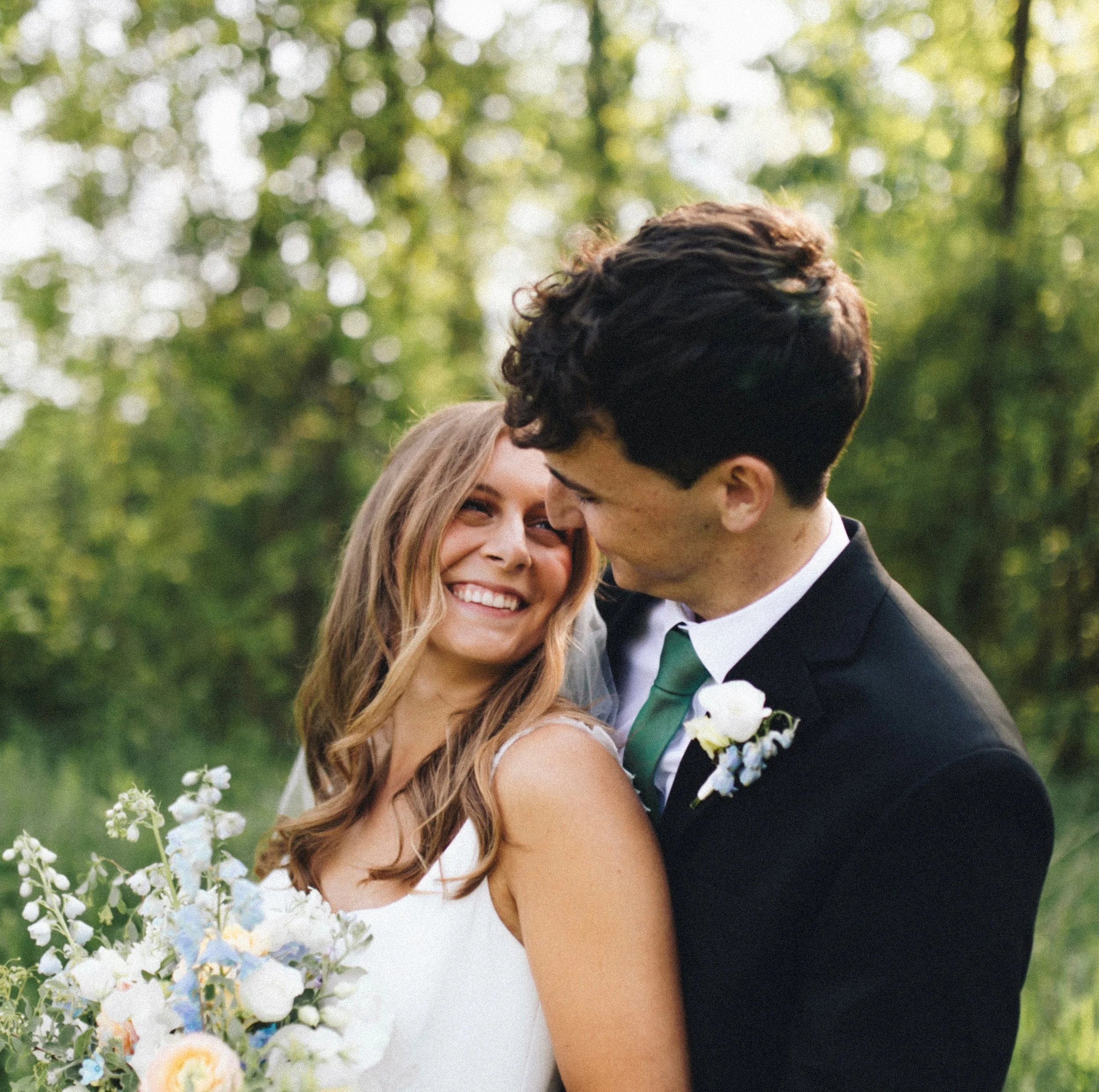 A happy couple in wedding attire sharing a close moment outdoors, with greenery and flowers surrounding them.