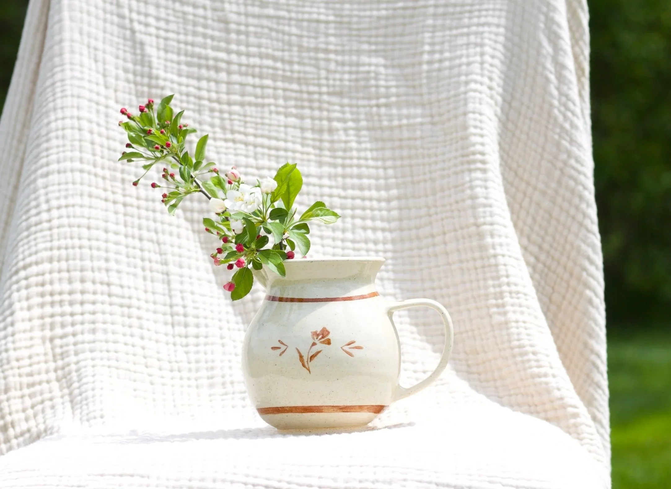 A white ceramic pitcher with orange-brown decorative designs, holding a small branch with white flowers, pink buds, and green leaves, in front of a textured white fabric and green background.