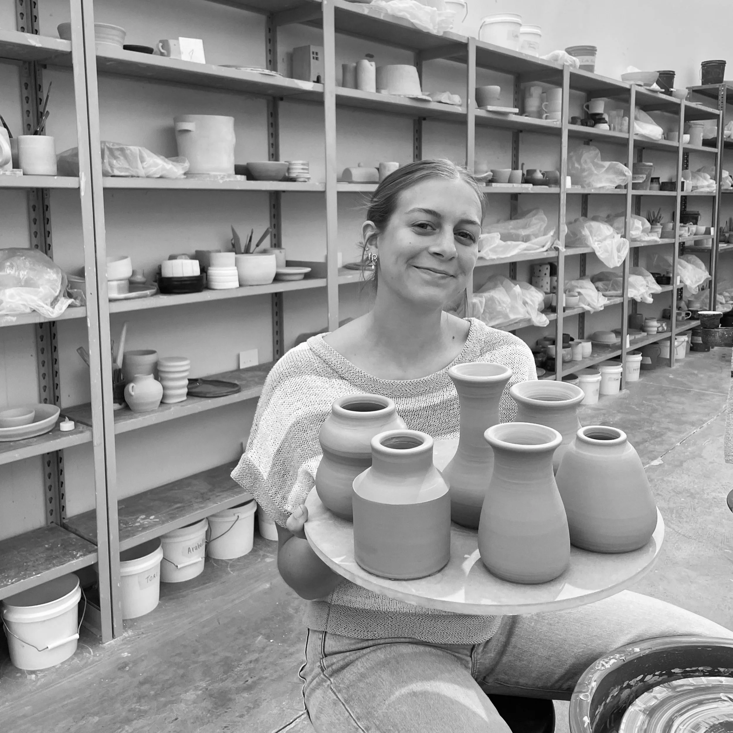 A woman smiling in a pottery studio. She is sitting at a table with various clay pots and sculptures. Behind her are shelves filled with bowls, cups, and other ceramic items, along with bags of clay and pottery tools.