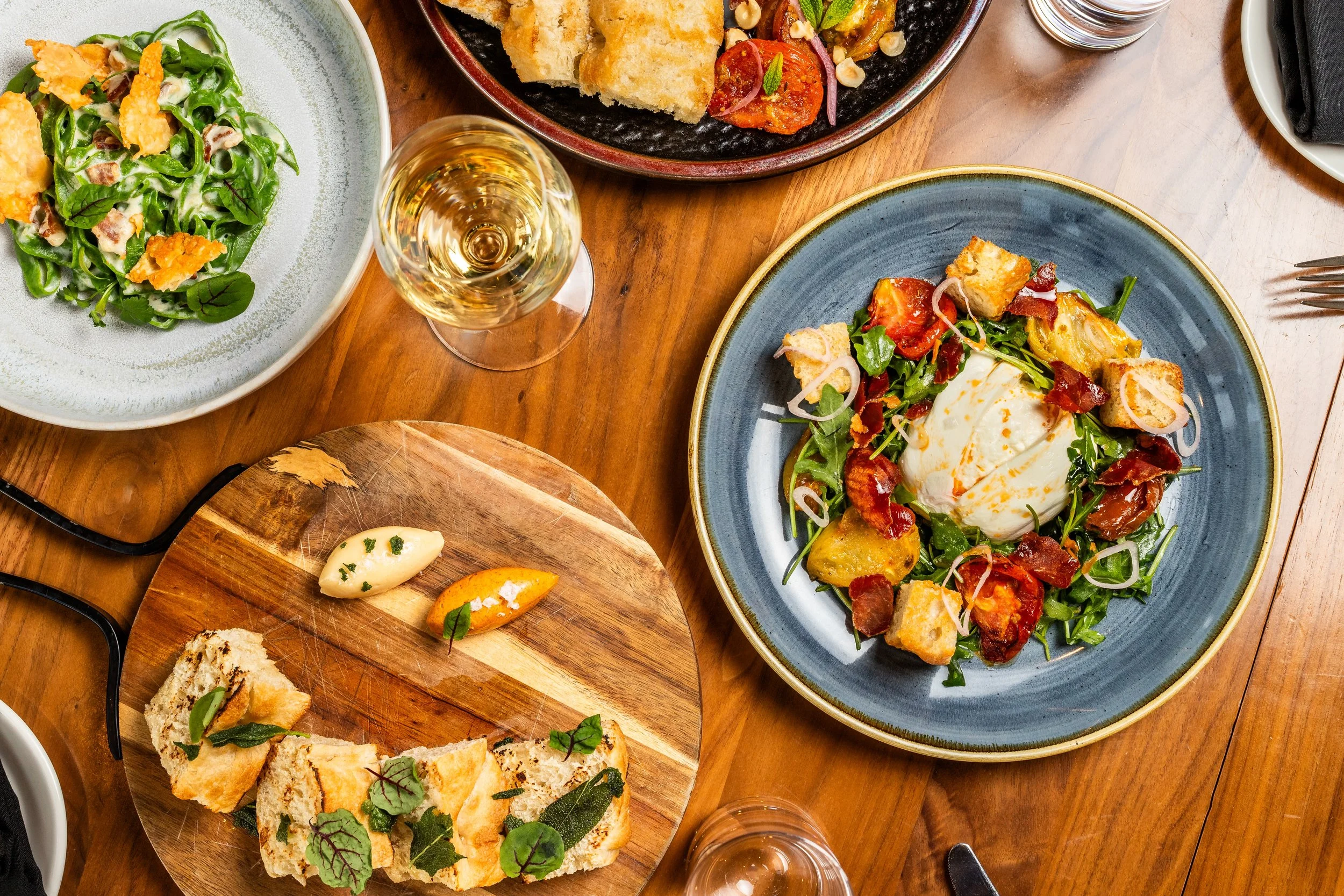 A dining table with a variety of dishes including a green salad, a main course with poached egg, roasted potatoes, and bacon on a blue plate, a rustic wooden board with bread slices, butter, and herbs, a glass of white wine, and other plates and uten