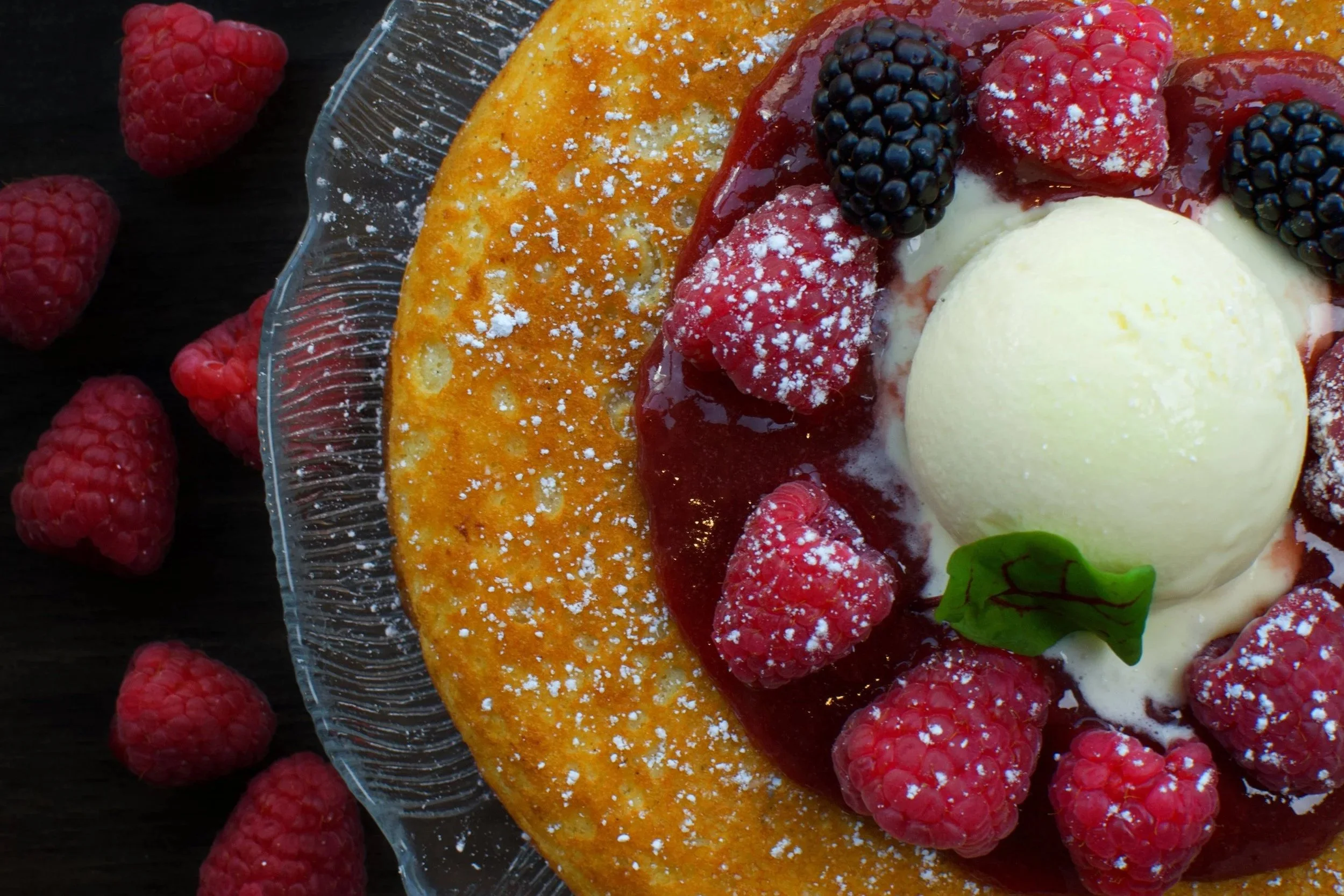 Close-up of a dessert with mixed berries, a scoop of vanilla ice cream, and a crispy crust, garnished with powdered sugar and a small green leaf, on a glass plate.