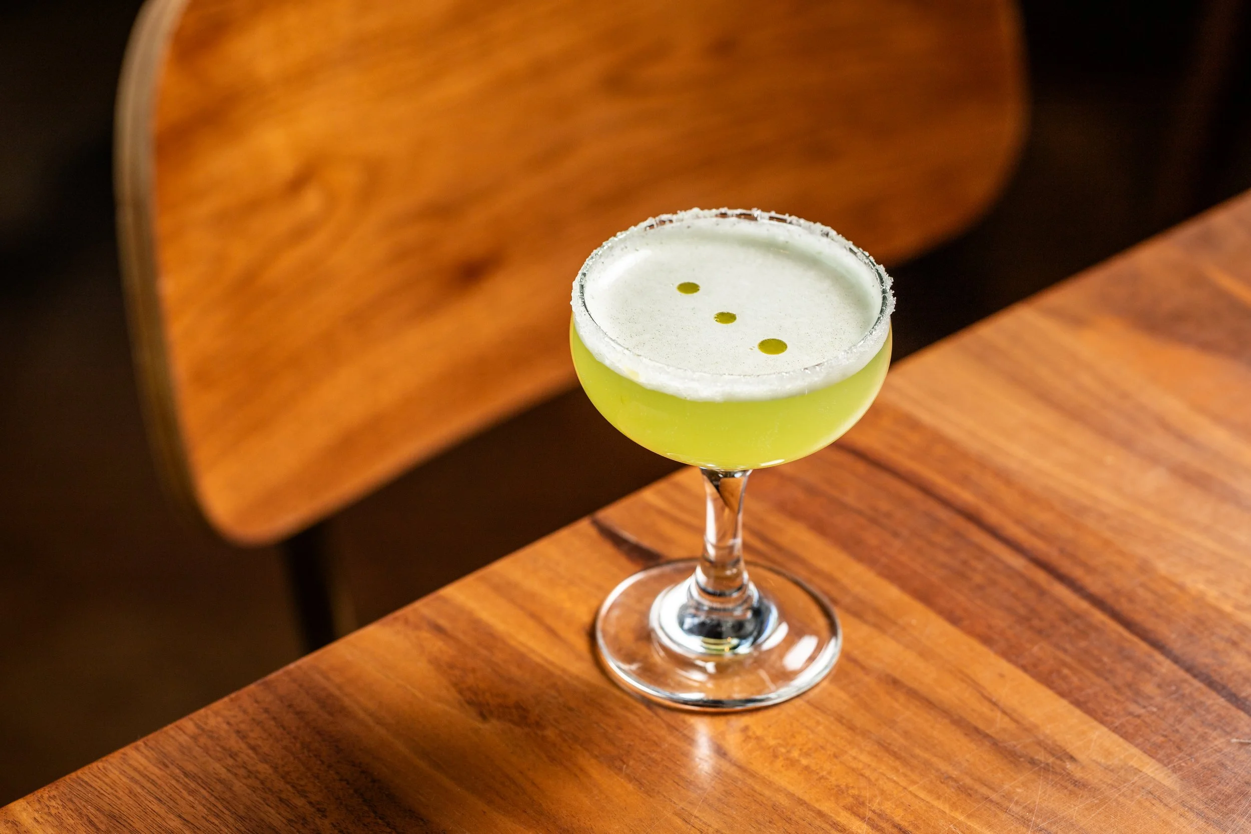 Green cocktail in a coupe glass with a salted rim and three small drops of liquid on top, placed on a wooden table with a wooden chair in the background.
