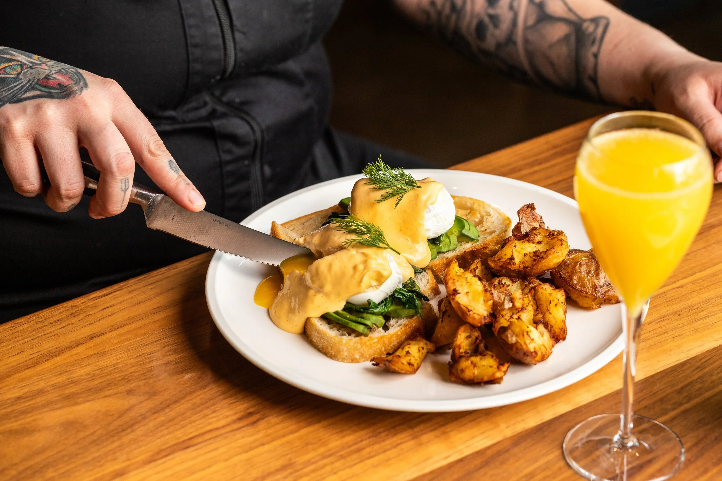 A person with tattoos is slicing eggs Benedict on a white plate that also has roasted potatoes and toast with greens and hollandaise sauce. There is a glass of orange juice.