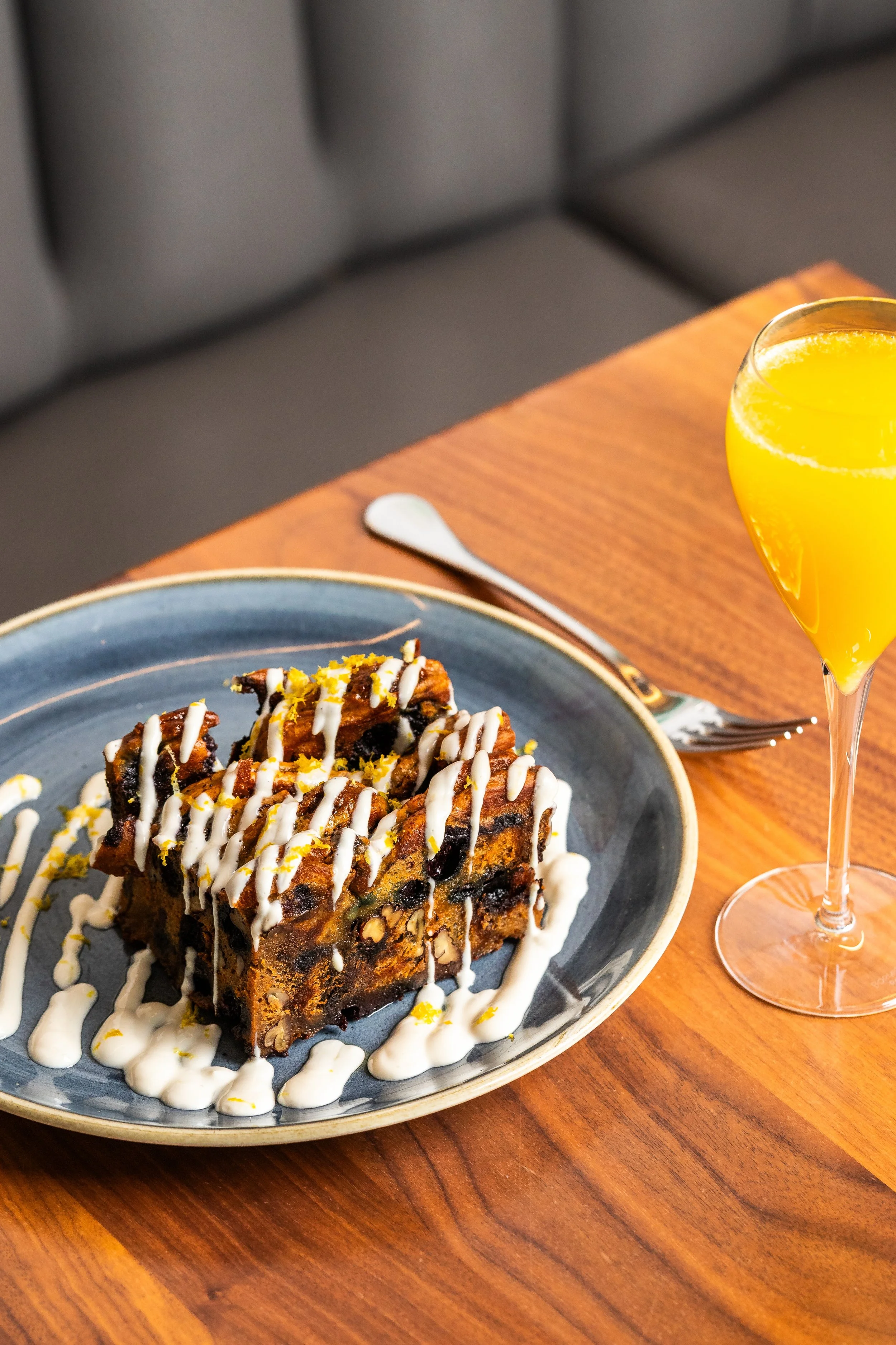A slice of decorated chocolate chip bread pudding topped with white icing drizzle on a blue plate, served with a glass of orange juice on a wooden table.