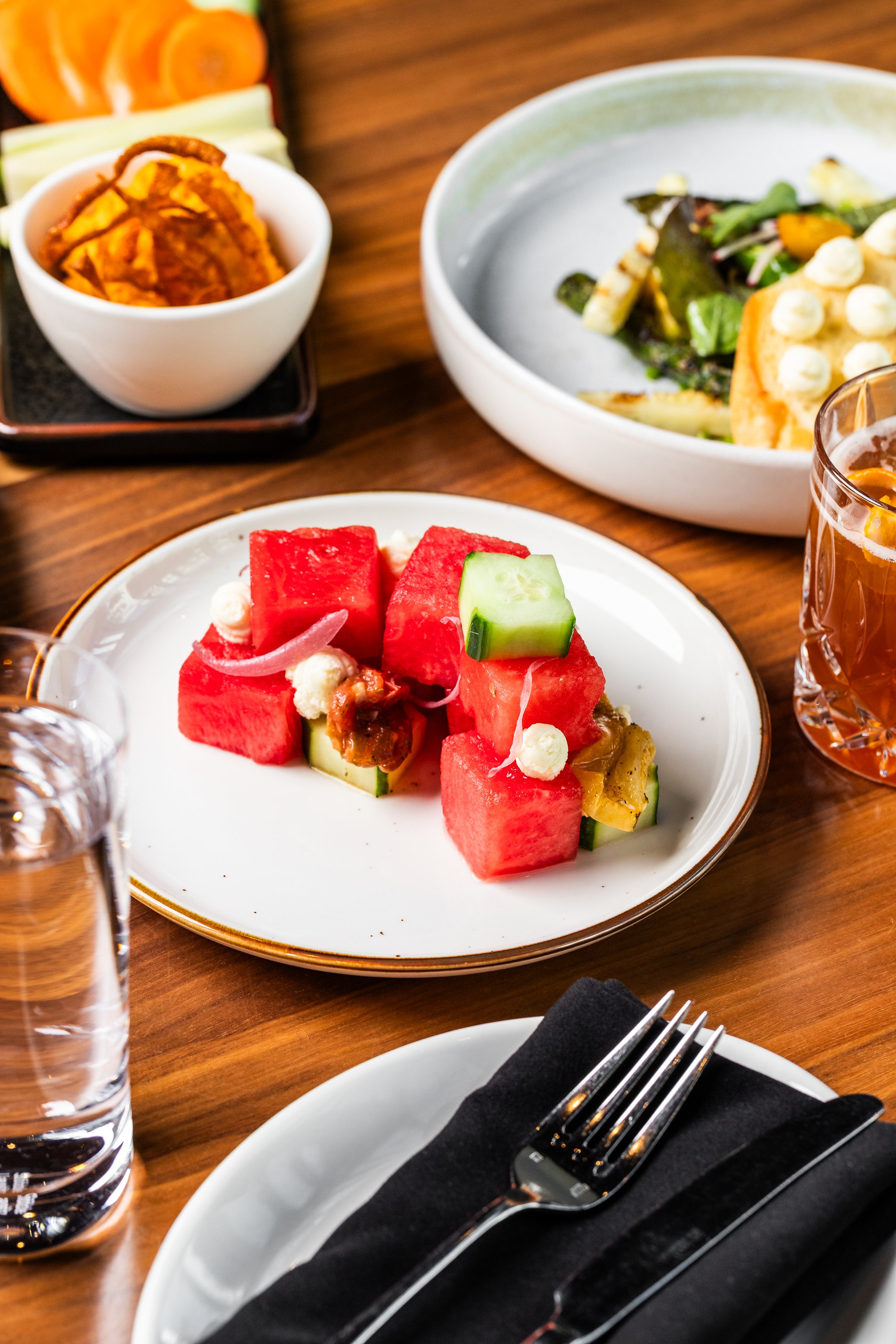 A white plate with watermelon cubes, cucumber slices, and small garnishes on a wooden table.