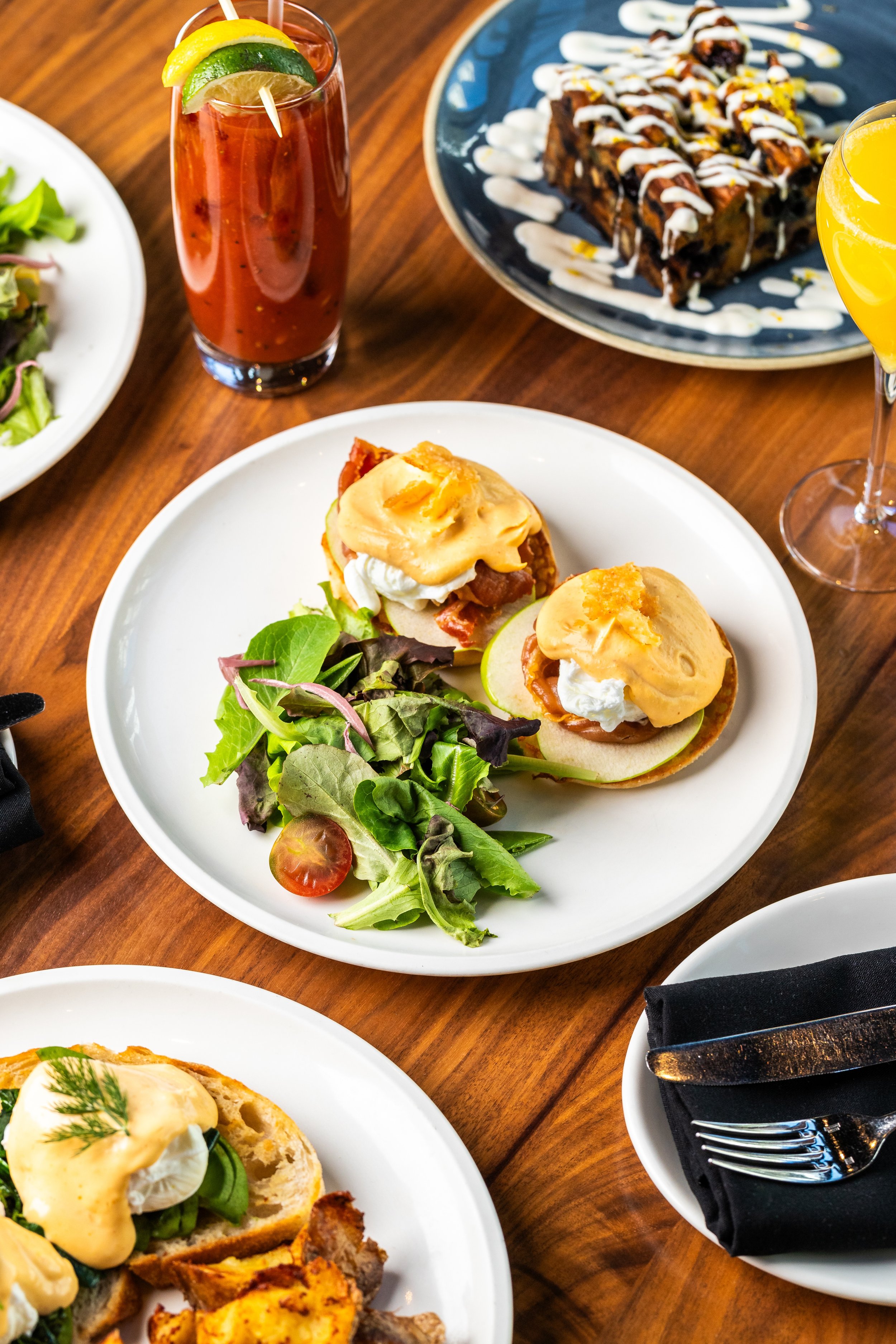 A plate with two open-faced sandwiches topped with poached eggs, bacon, and hollandaise sauce, served with a side salad. Nearby are other dishes including a slice of savory bread with eggs, and two beverages; a Bloody Mary and a mimosa.