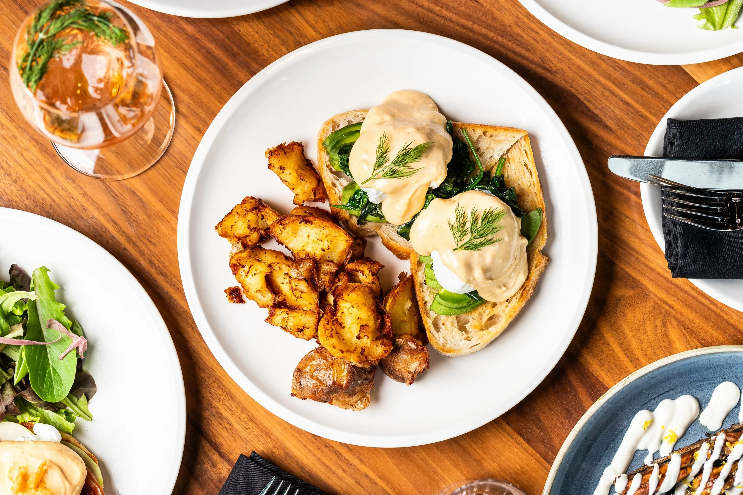 Plate with open-faced sandwiches topped with poached eggs, greens, and hollandaise sauce, alongside roasted potatoes; glass of iced tea and other dishes on a wooden table.