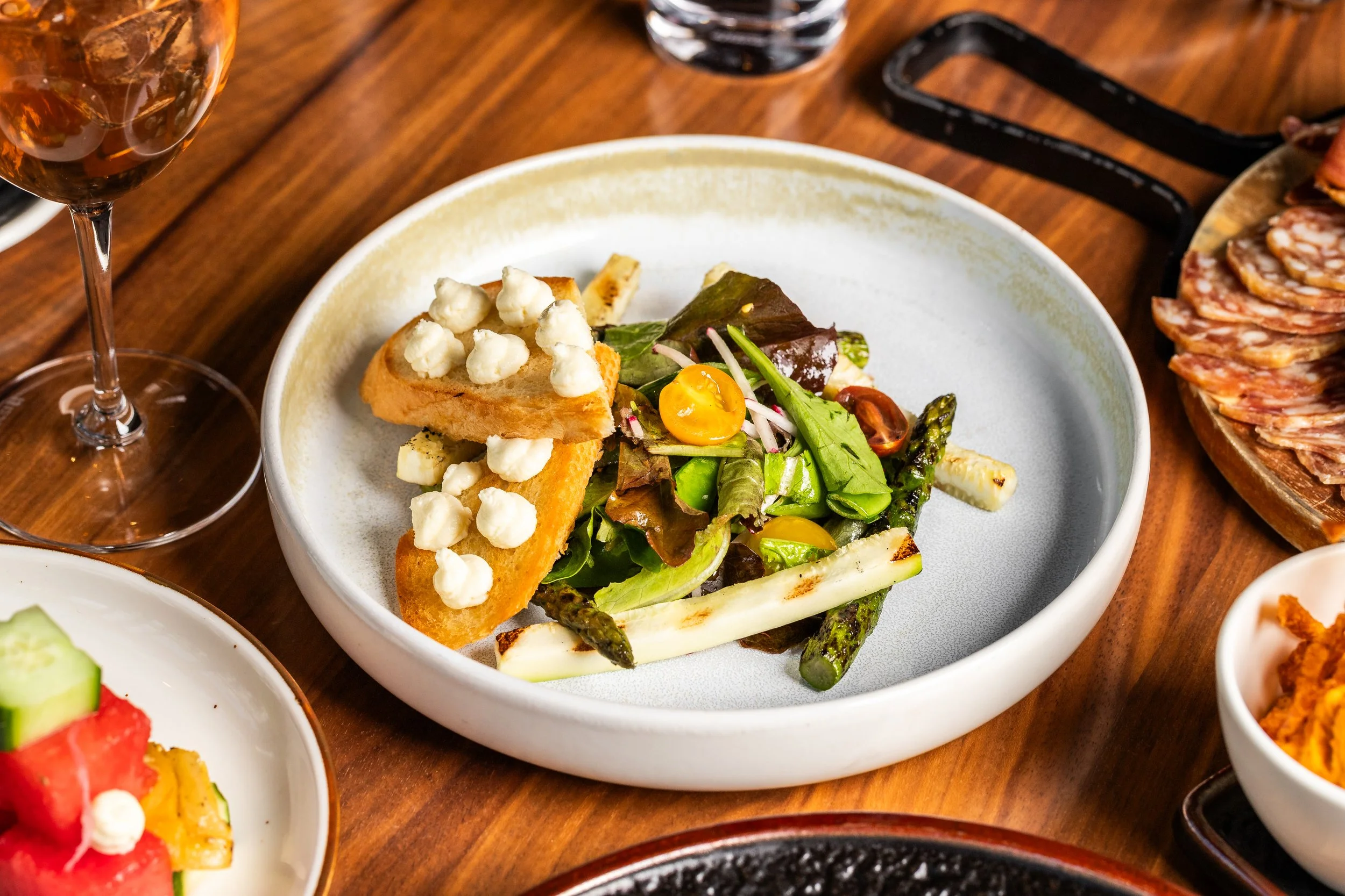 A white bowl with a mixed green salad containing cherry tomatoes, grilled asparagus, and croutons topped with cheese