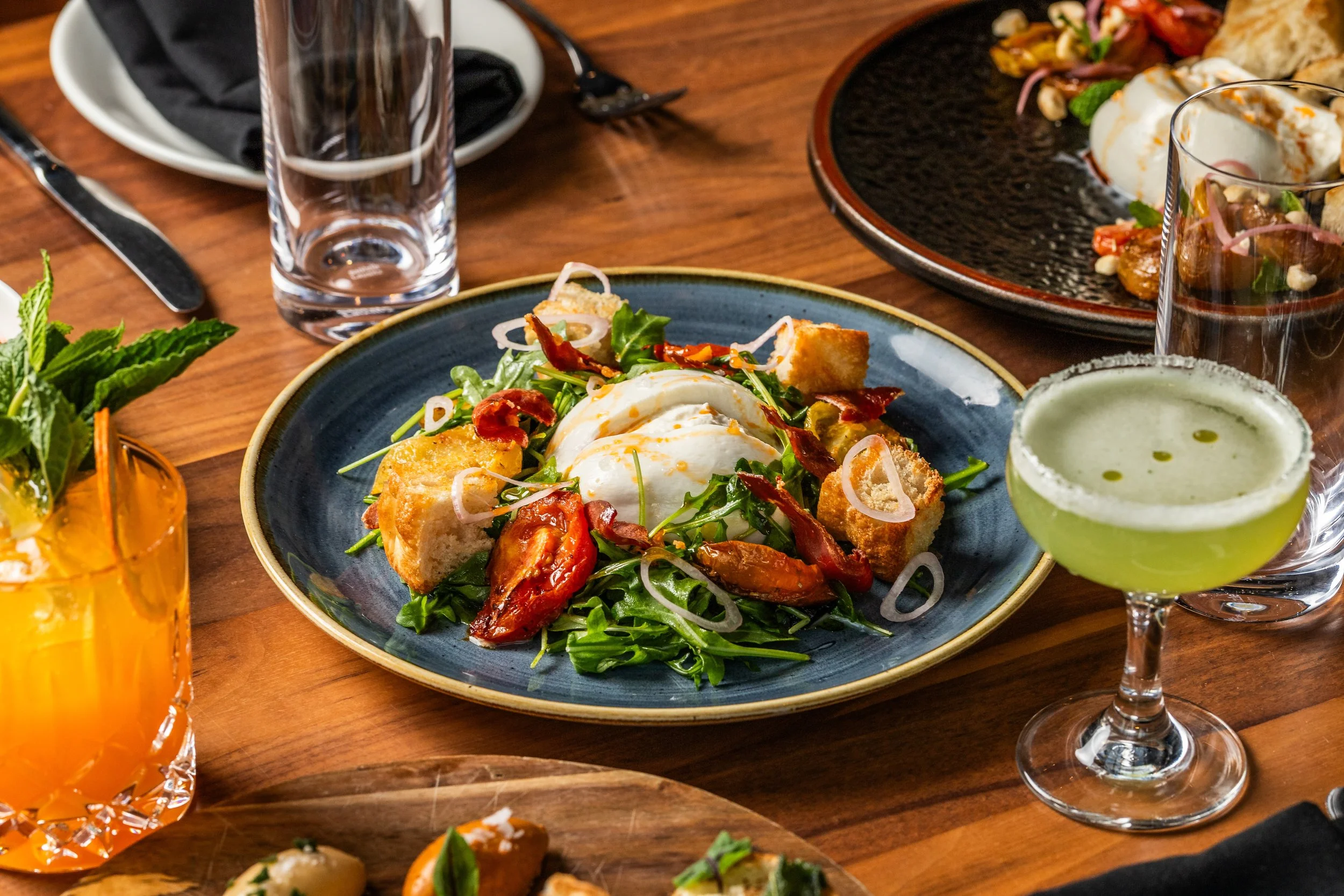 A colorful salad with greens, cherry tomatoes, bacon bits, onion slices, and a poached egg on a blue plate, accompanied by a green cocktail, an orange beverage, and empty glasses on a wooden table.