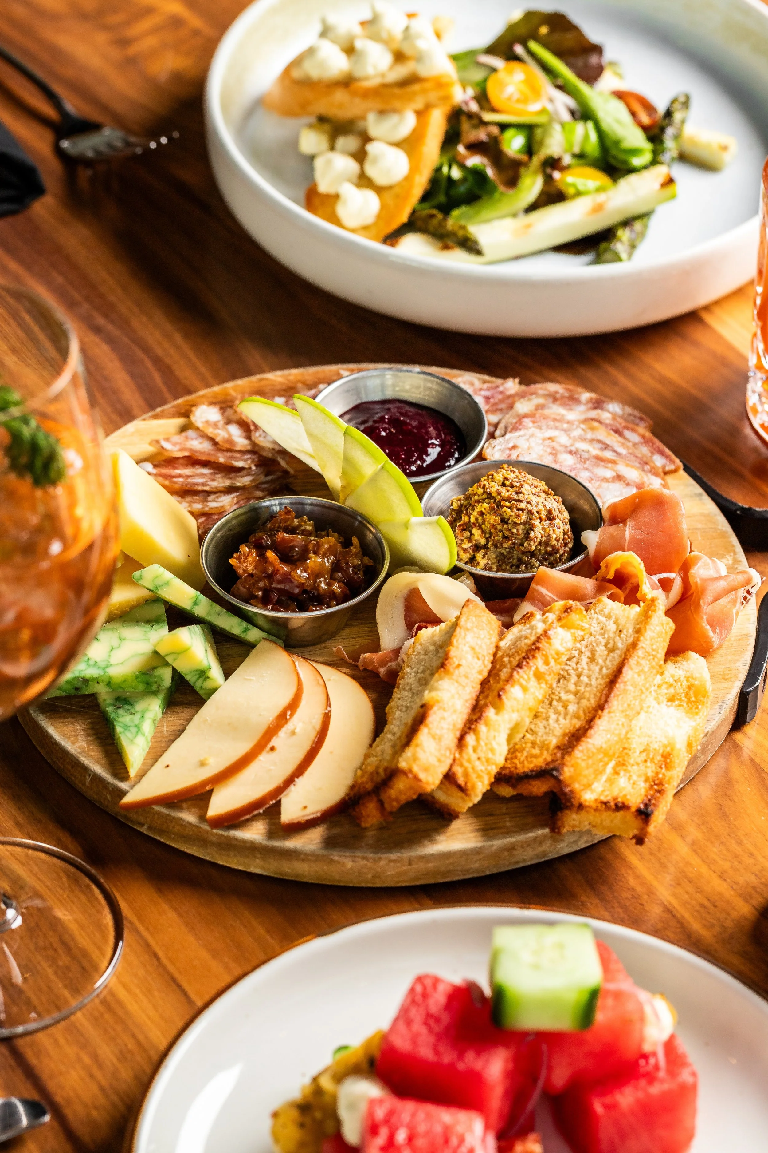 Charcuterie board with assorted cheeses, cured meats, fruit, and dips, set on a wooden table. In the background, a salad with greens and grilled vegetables. Also visible are a glass of rosé wine and a plate of watermelon and other fruits.