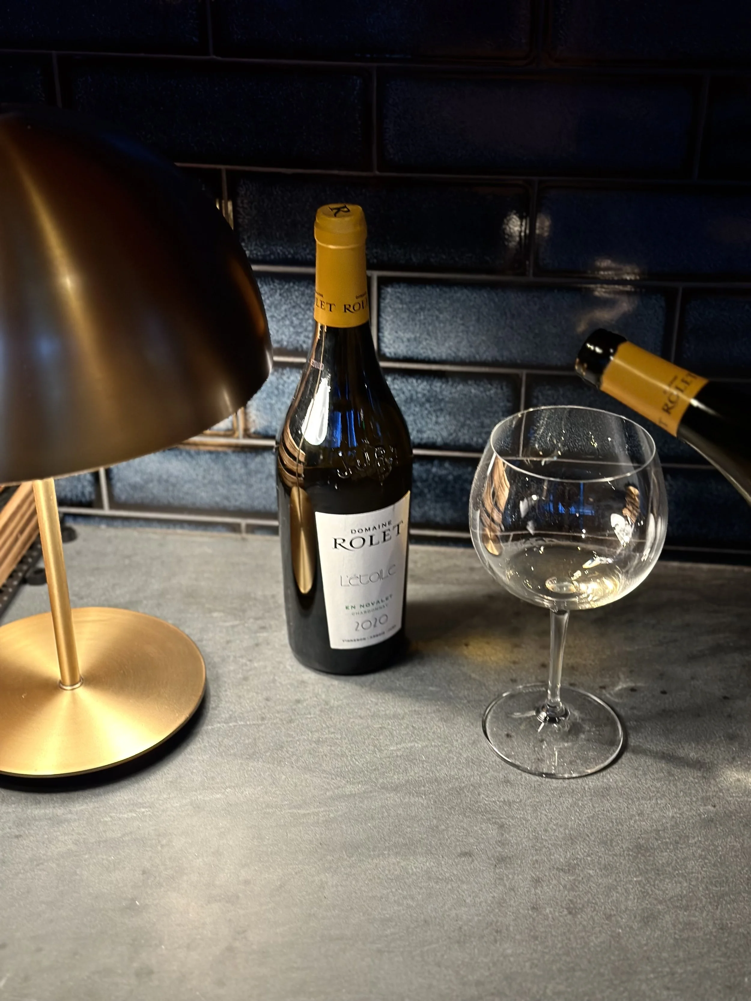 A bottle of Domaine Rolet Le Voluc 2010 wine with a yellow capsule, an empty wine glass, and a gold table lamp on a gray surface against a dark tiled wall.
