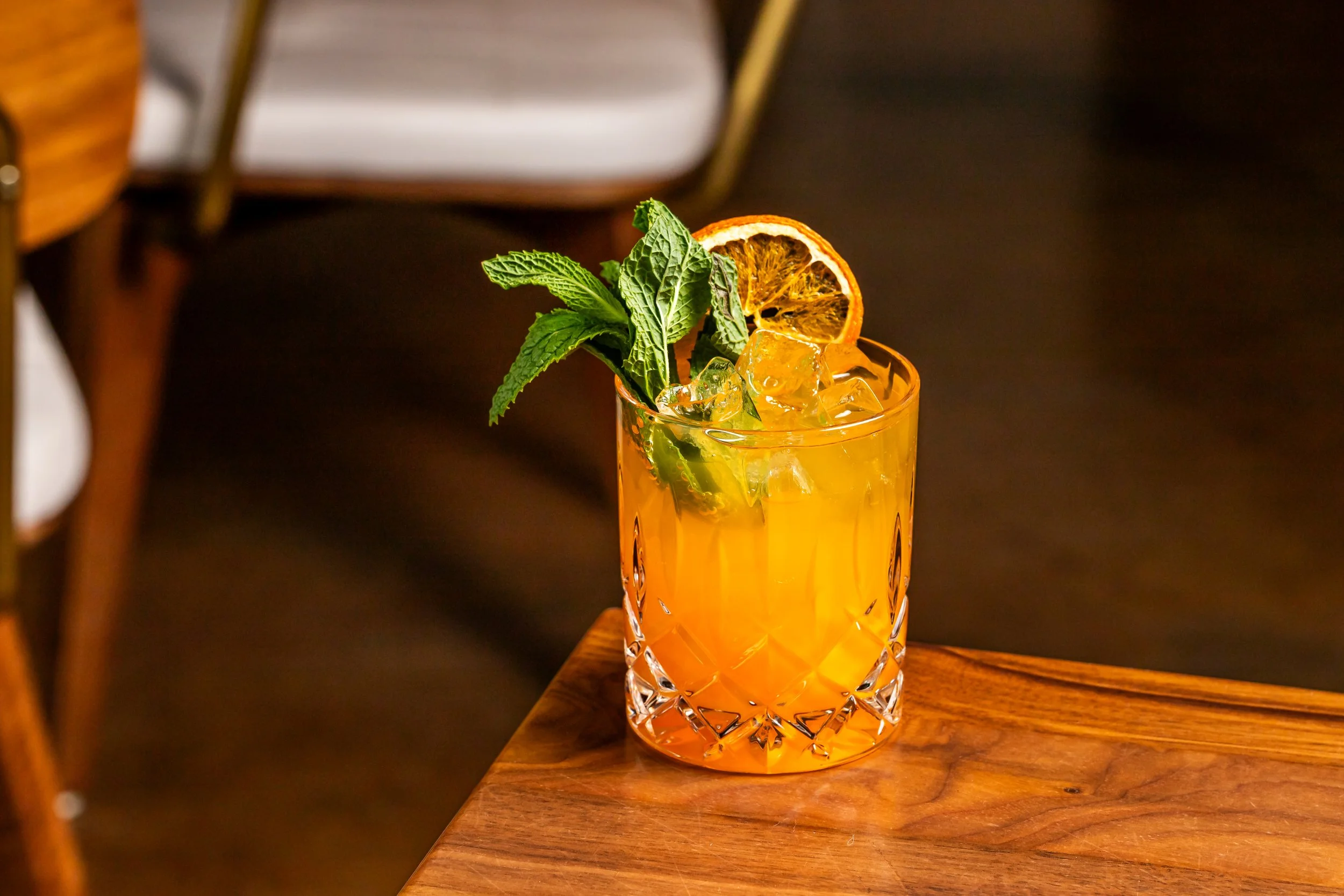 A glass of orange cocktail garnished with mint leaves and an orange slice, served with ice, on a wooden table.