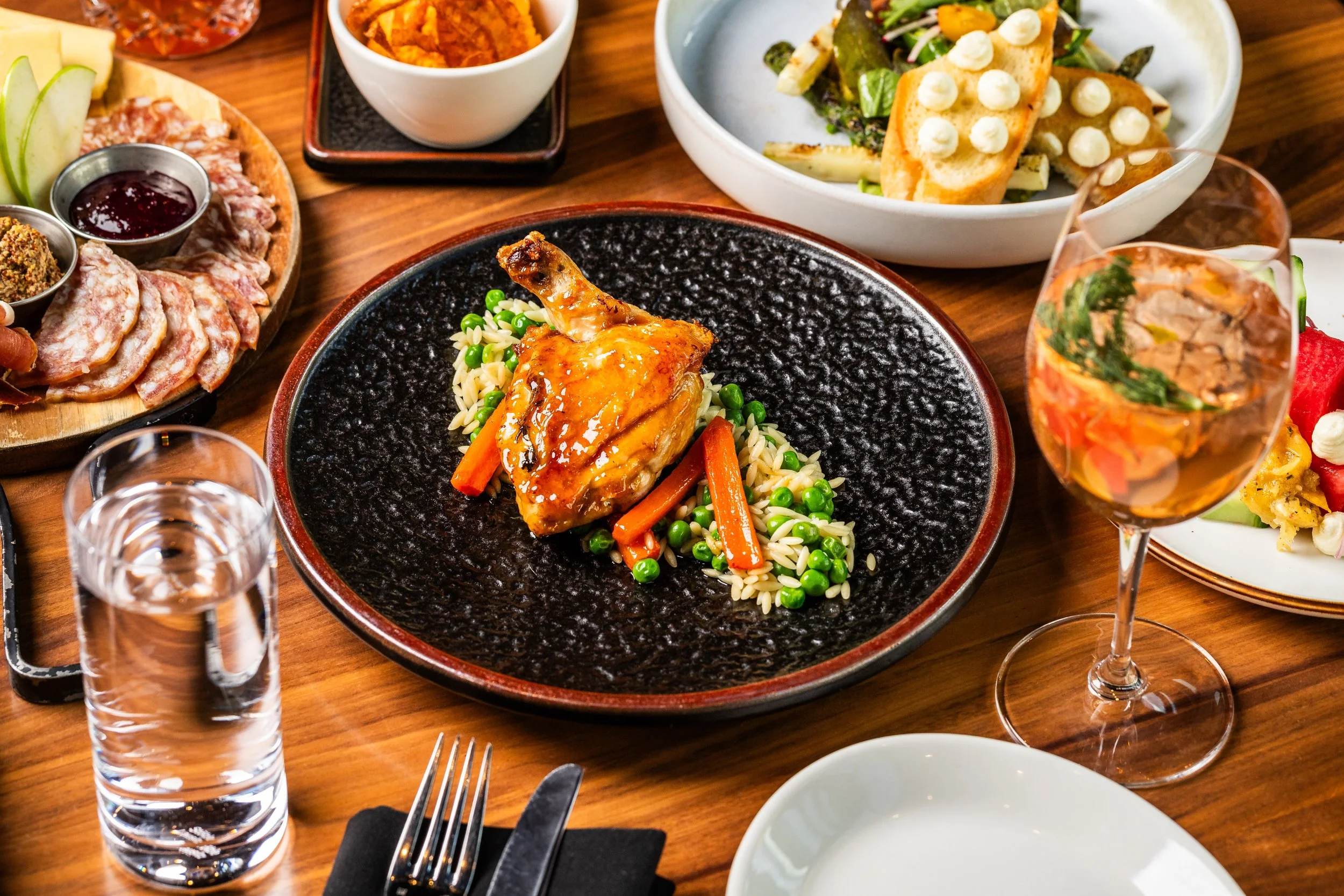 A dinner table with a cooked piece of chicken with vegetables, a glass of water, a glass of wine, and various side dishes including salad, bread with toppings, and dessert.
