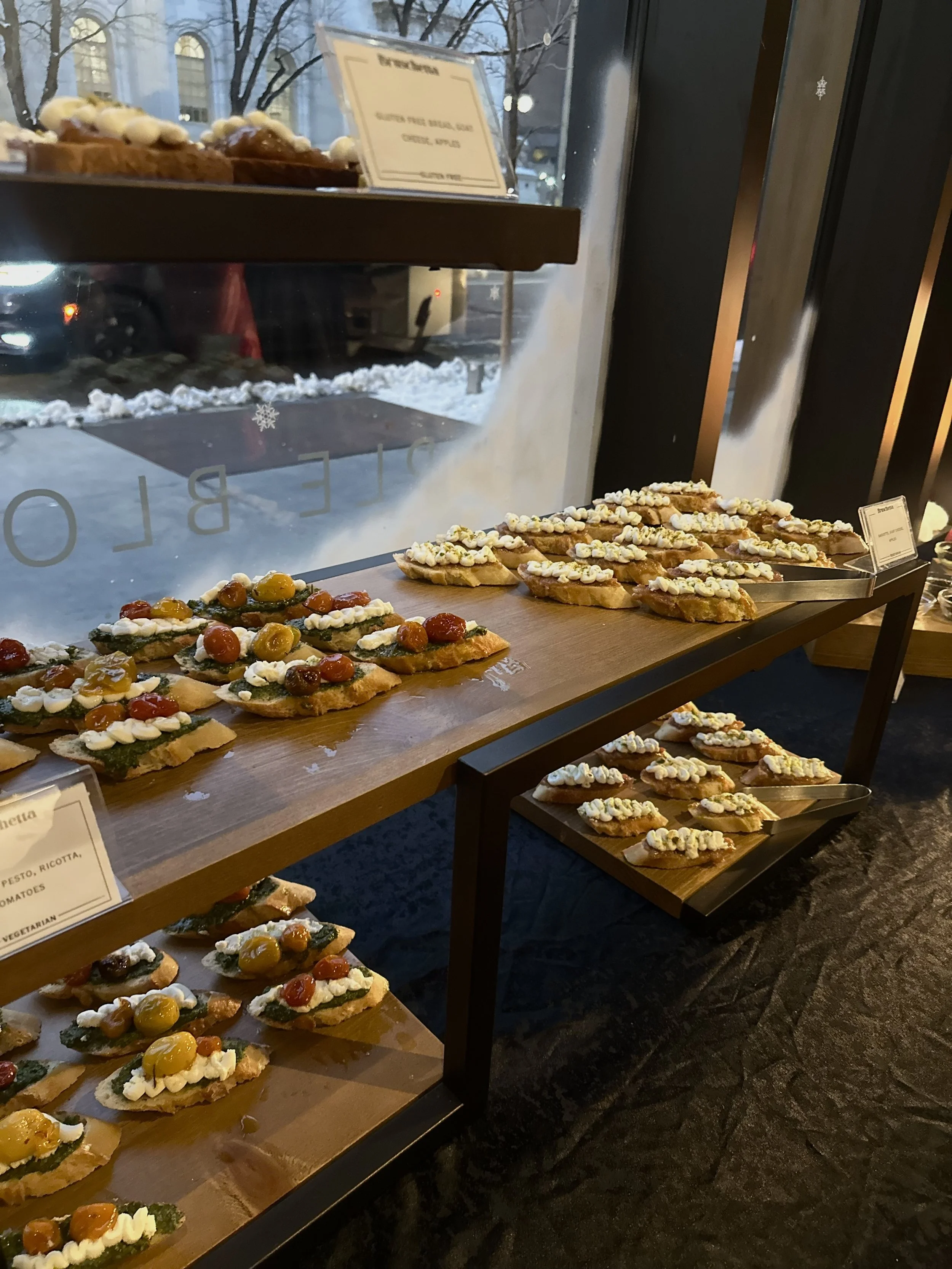 Assorted appetizers on wooden trays display. Topped with whipped cheese, cherry tomatoes, and pesto. Inside a restaurant near a window, snow outside, evening setting.