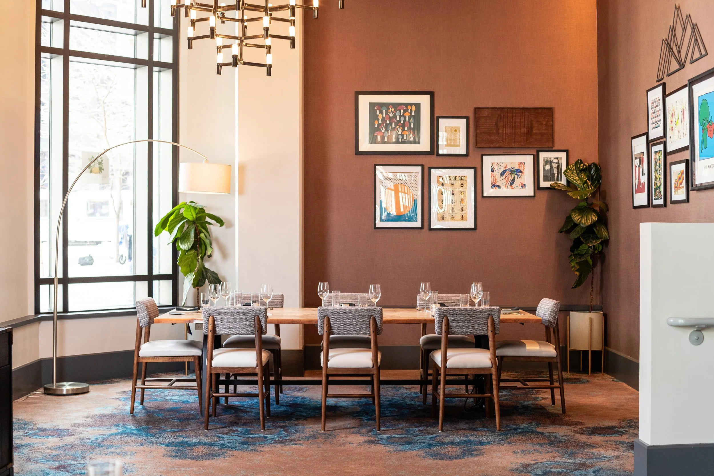 A dining area with a wooden table, ten chairs with beige cushions, wine glasses set on the table, large window with black frame, patterned carpet, framed art on a pink accent wall, a tall green plant near a white floor lamp, and ceiling chandelier.