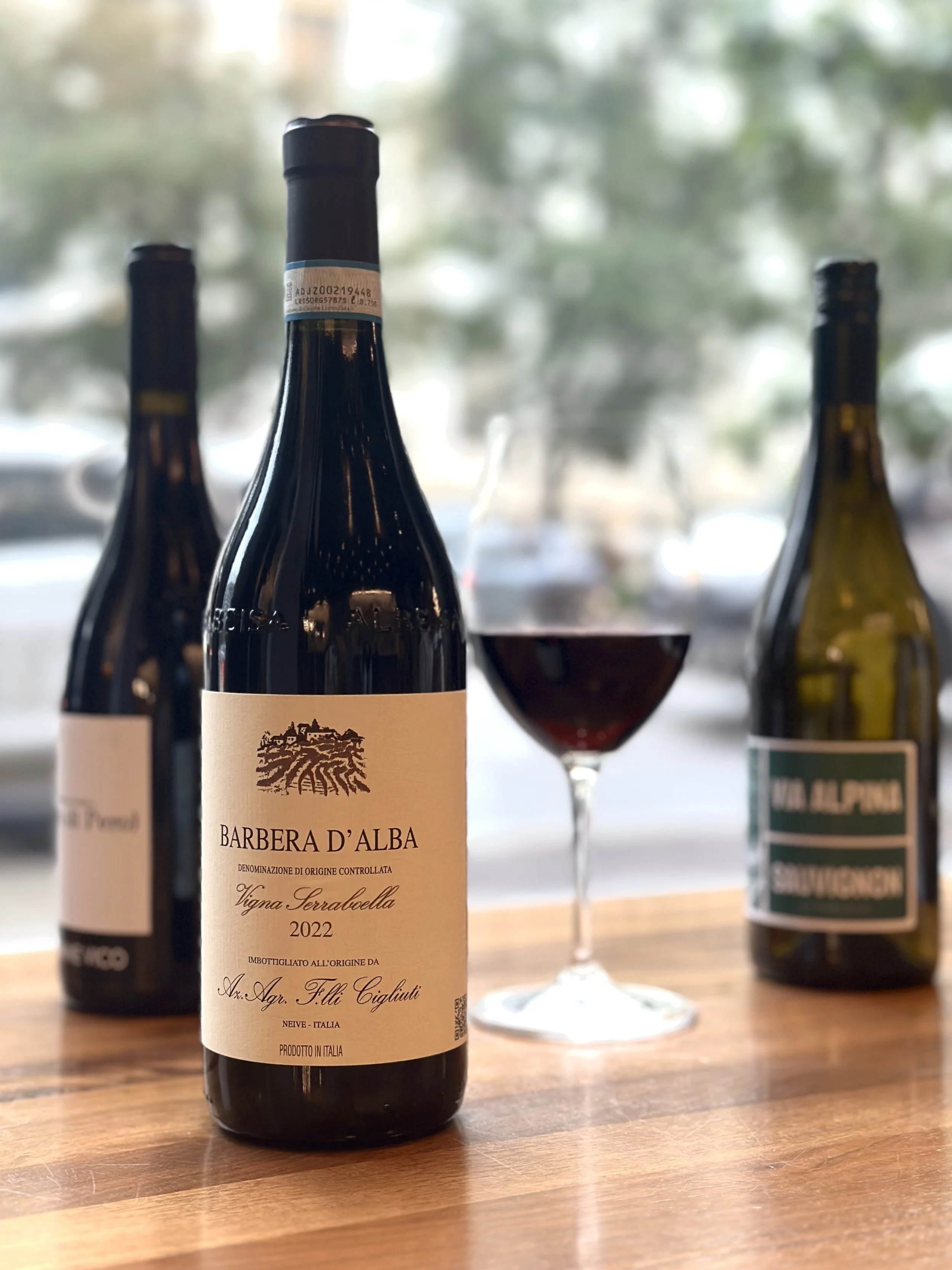 A bottle of Barbera D'Alba wine, a glass of red wine, and two other bottles in the background on a wooden table.