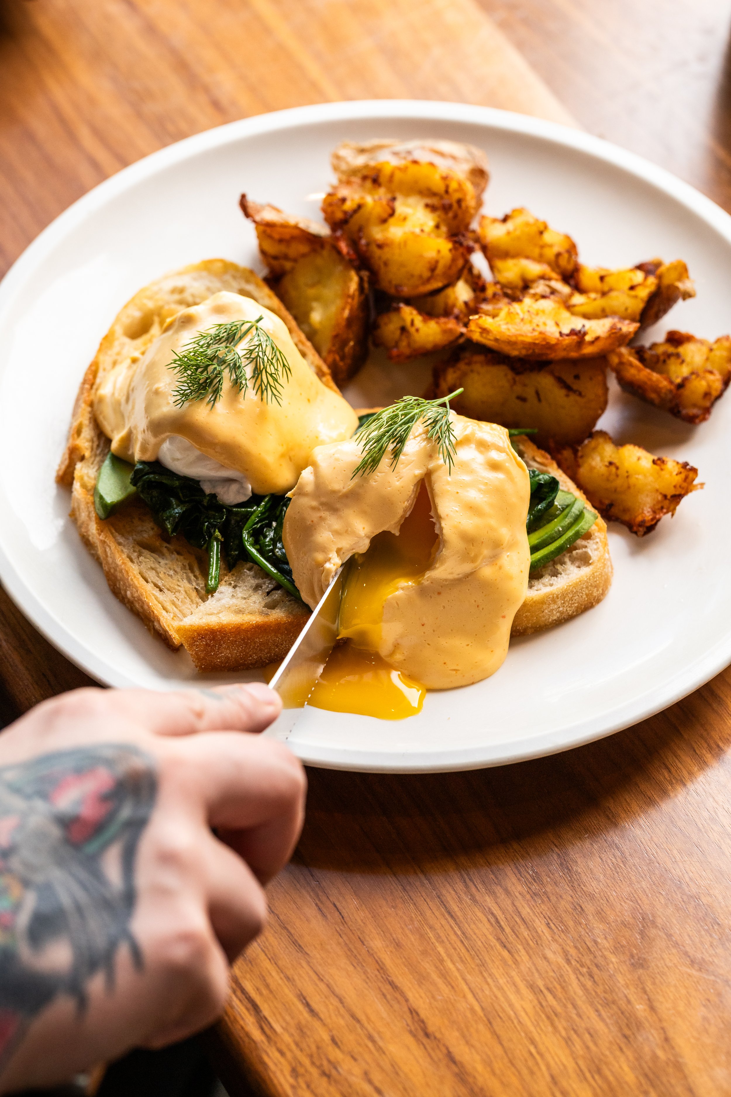 A plate with eggs Benedict, spinach, avocado, roasted potatoes, and grilled chicken, with a tattooed hand cutting into the eggs.
