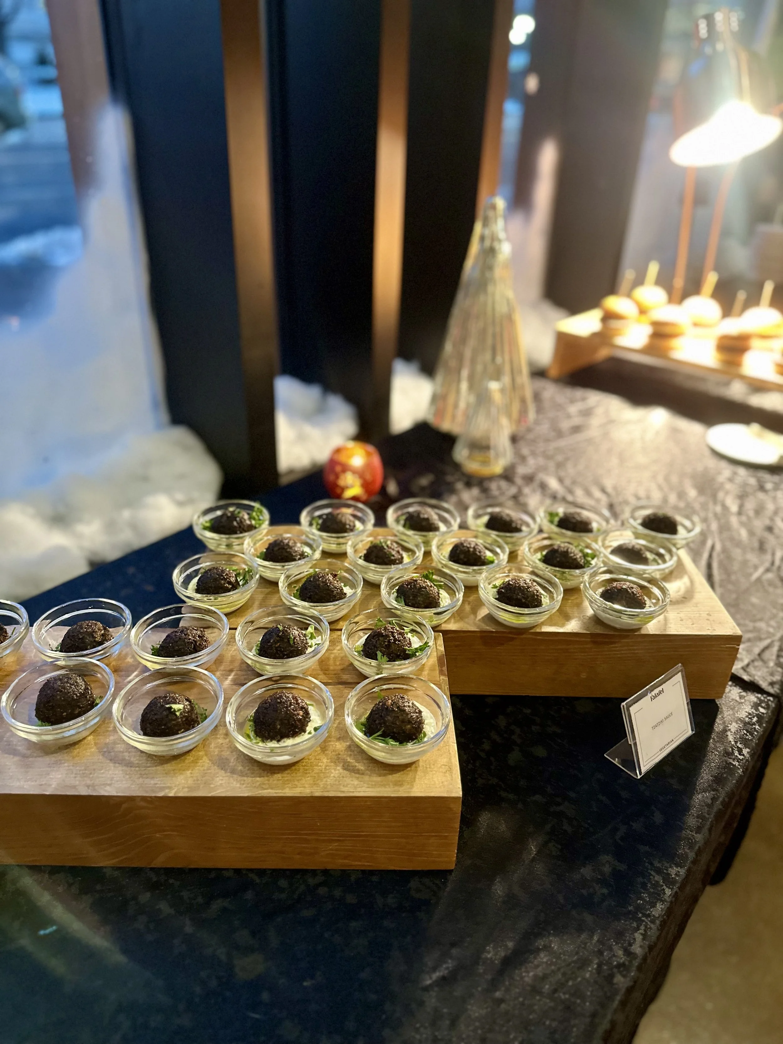 Small glass bowls with dark food items, possibly meatballs or truffles, garnished with herbs, arranged on wooden boards at a table.