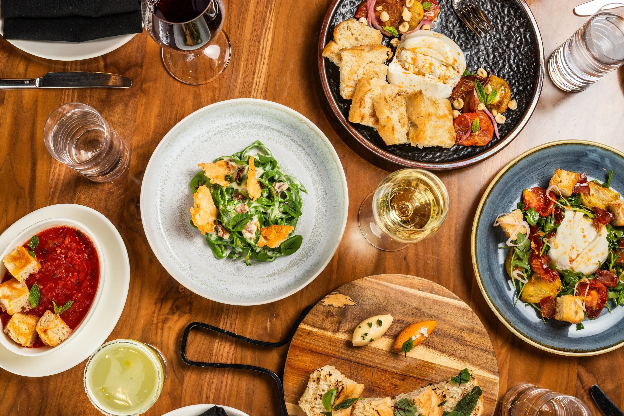 A top-down view of a wooden table with various dishes including salads, bread, wine, and water glasses.