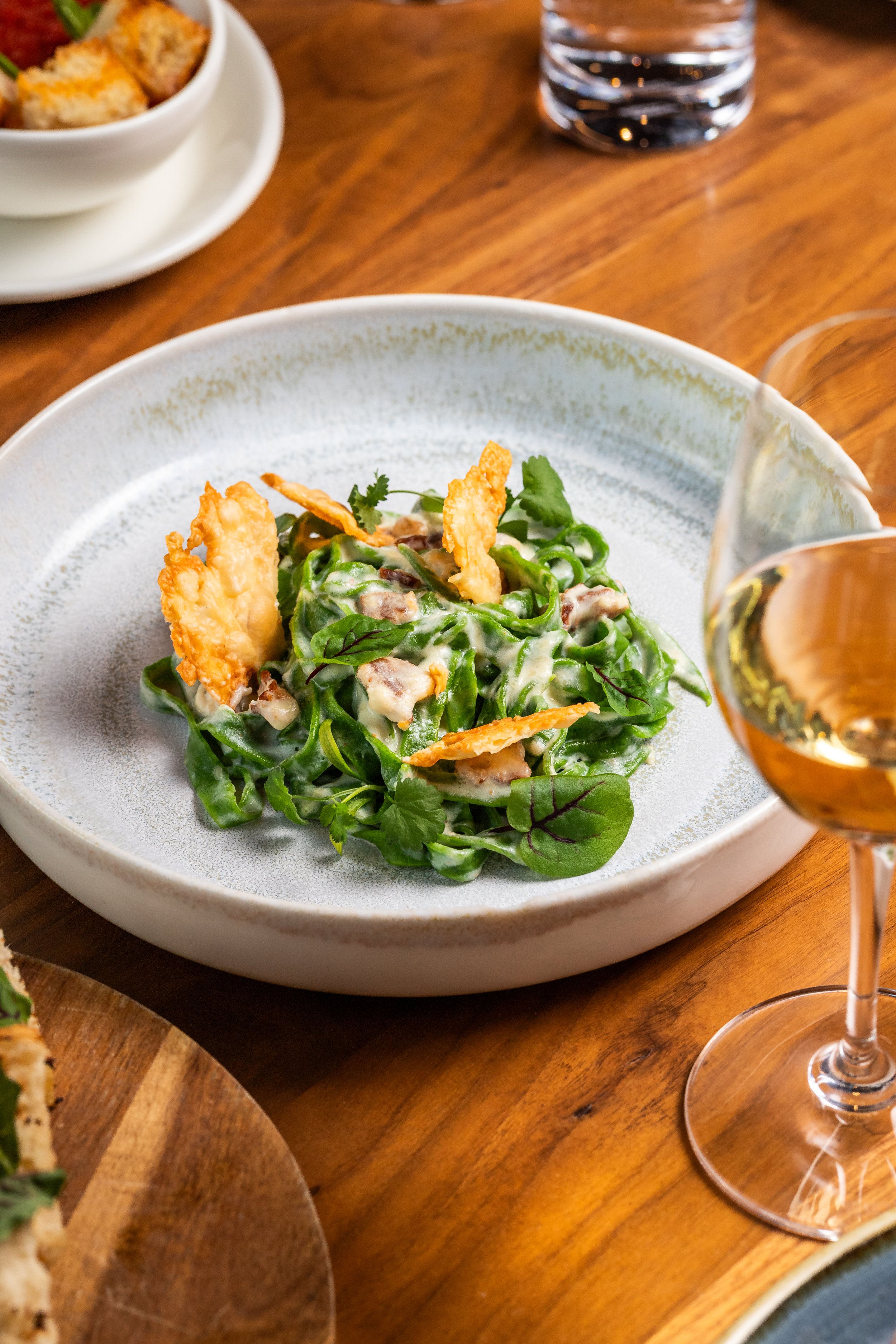 A salad with greens, creamy dressing, and crispy toppings served on a white plate, with a glass of white wine to the right.