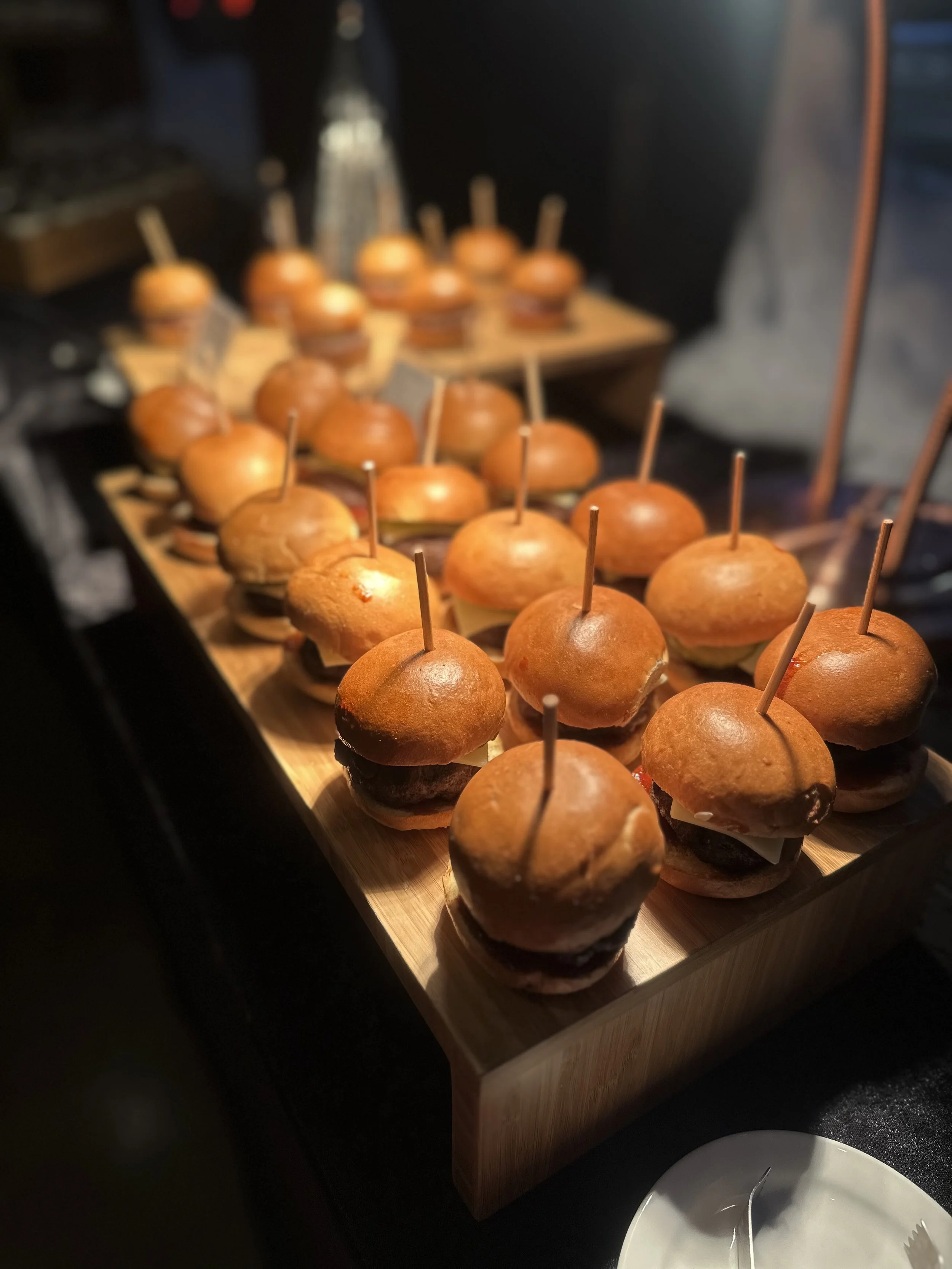 Mini cheeseburgers with buns and toothpicks on a wooden platter at a food event.