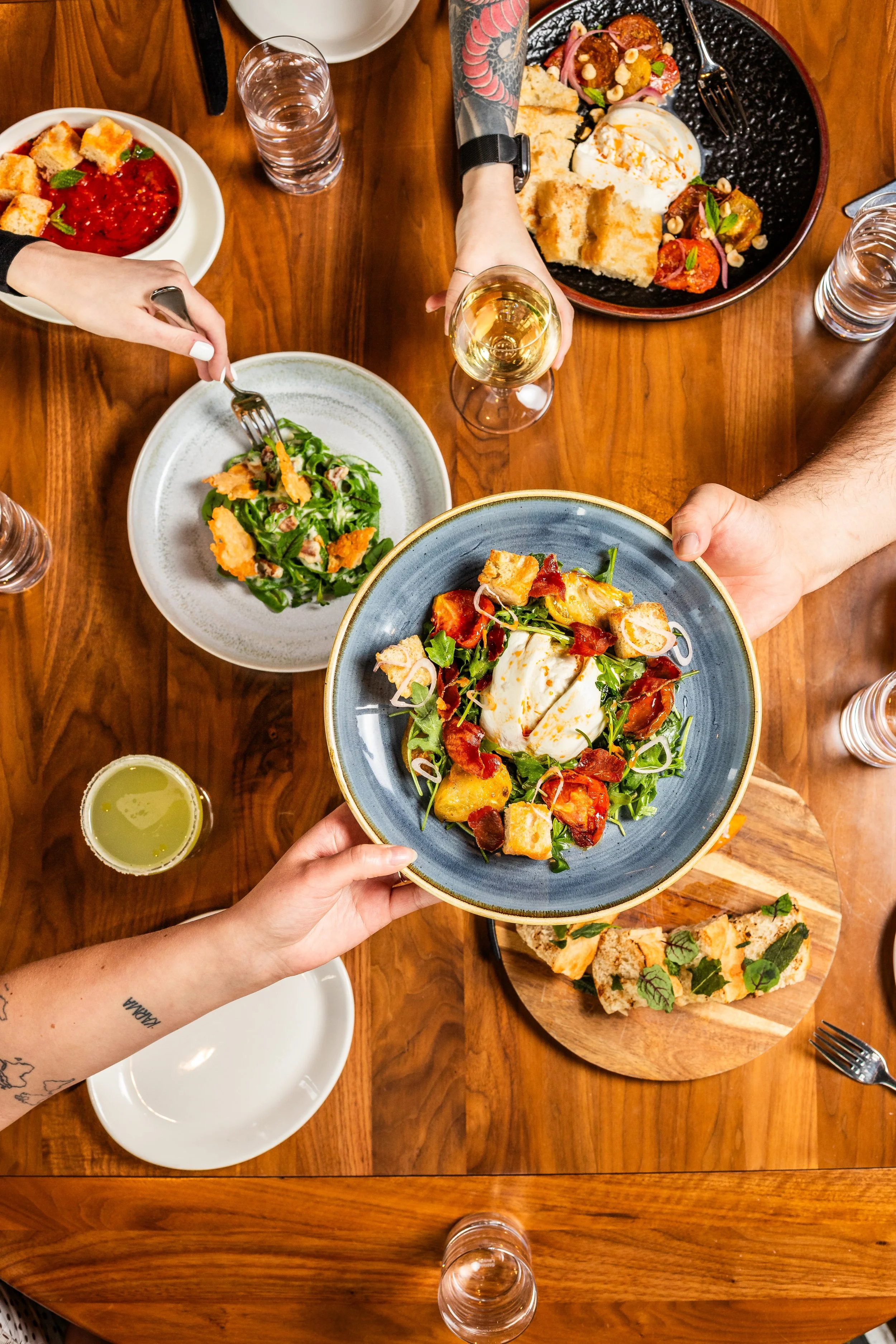 People sharing a meal of salads, bread, and drinks at a wooden table, with one person holding a plate with a salad topped with burrata cheese.