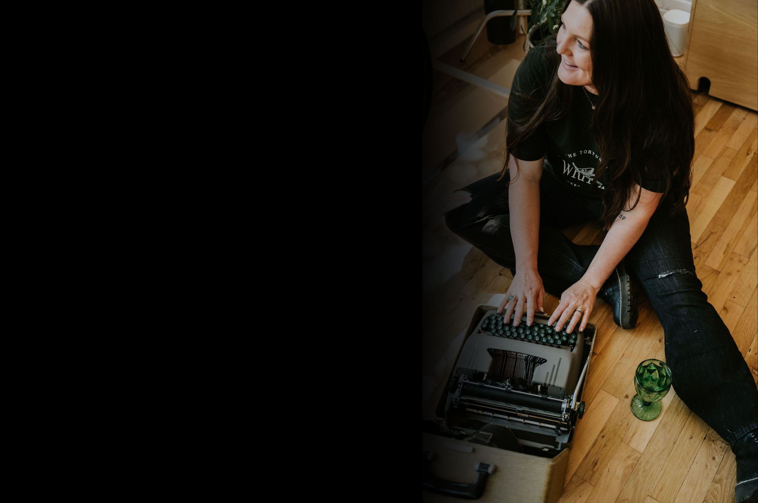 A woman with long dark hair is sitting on the wooden floor, smiling, and typing on an old-fashioned typewriter. She is wearing a black T-shirt, ripped jeans, and black boots. There is a green glass vase and a potted plant in the background.