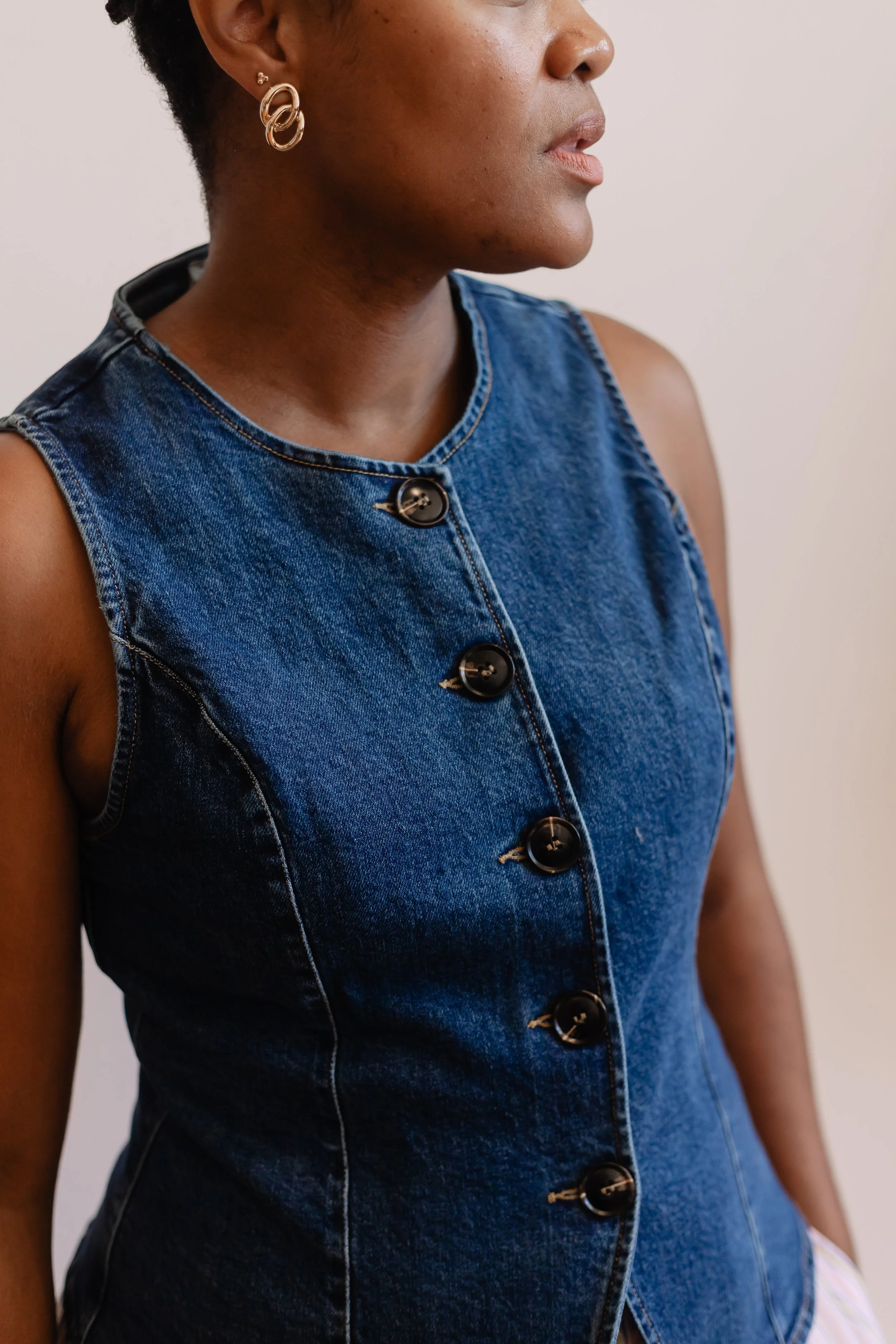 A woman wearing a sleeveless denim dress with black buttons, gold hoop earrings, and a small earring, standing against a plain background.