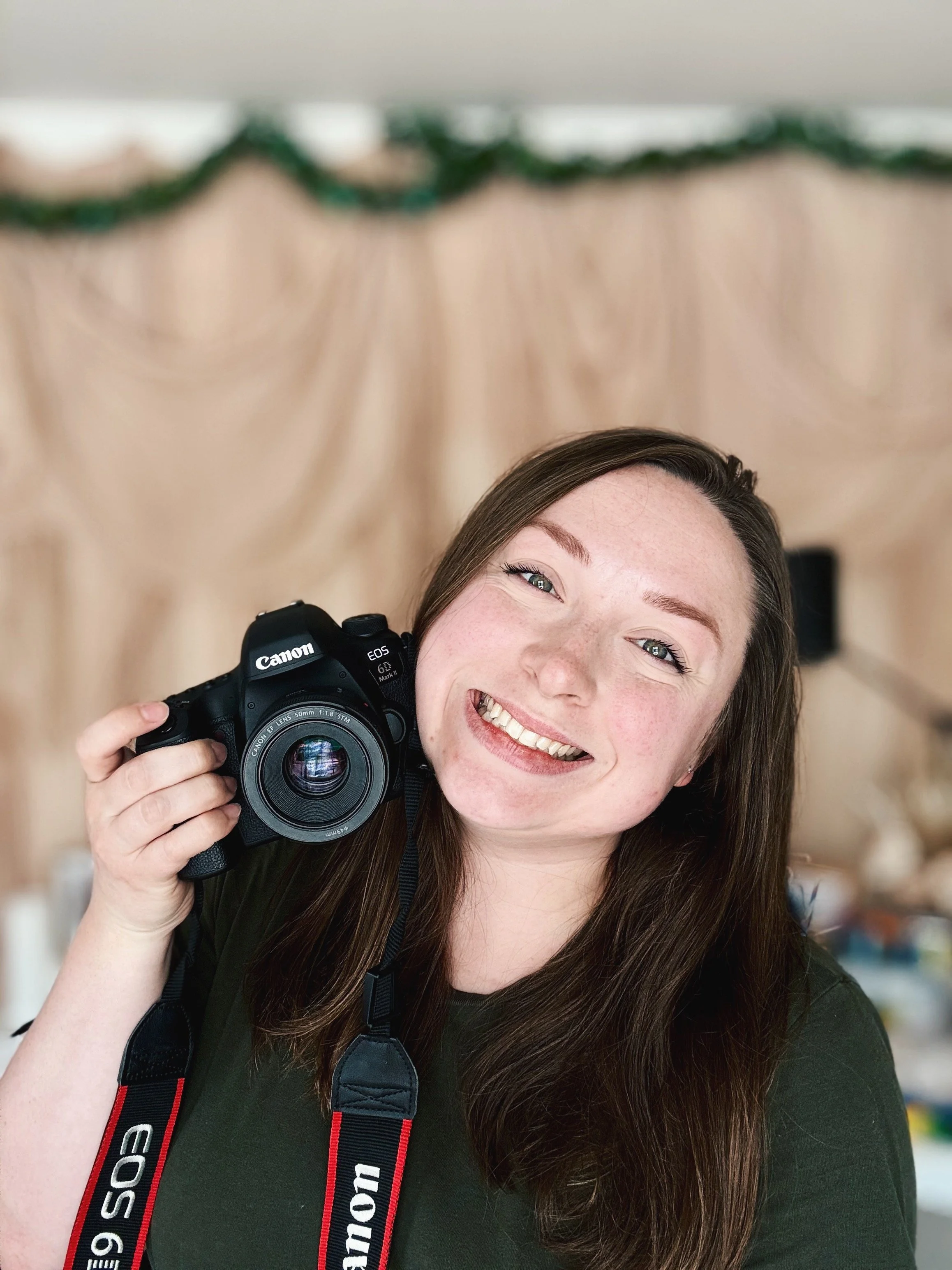 A woman with long brown hair smiling while holding a Canon camera with the strap around her neck, in front of a beige background with green garland.