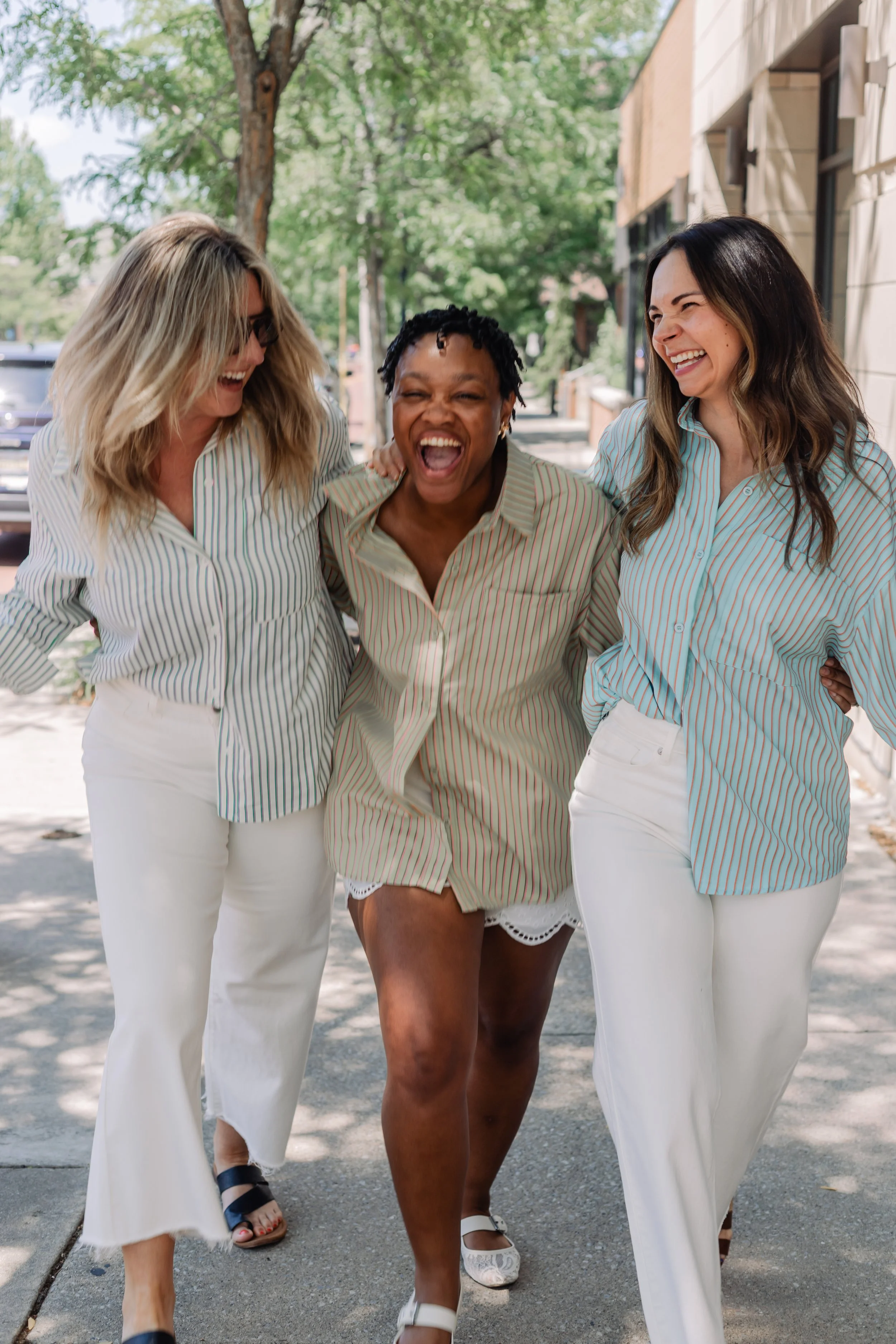 Three women smiling and laughing while walking arm-in-arm on a sunny street.