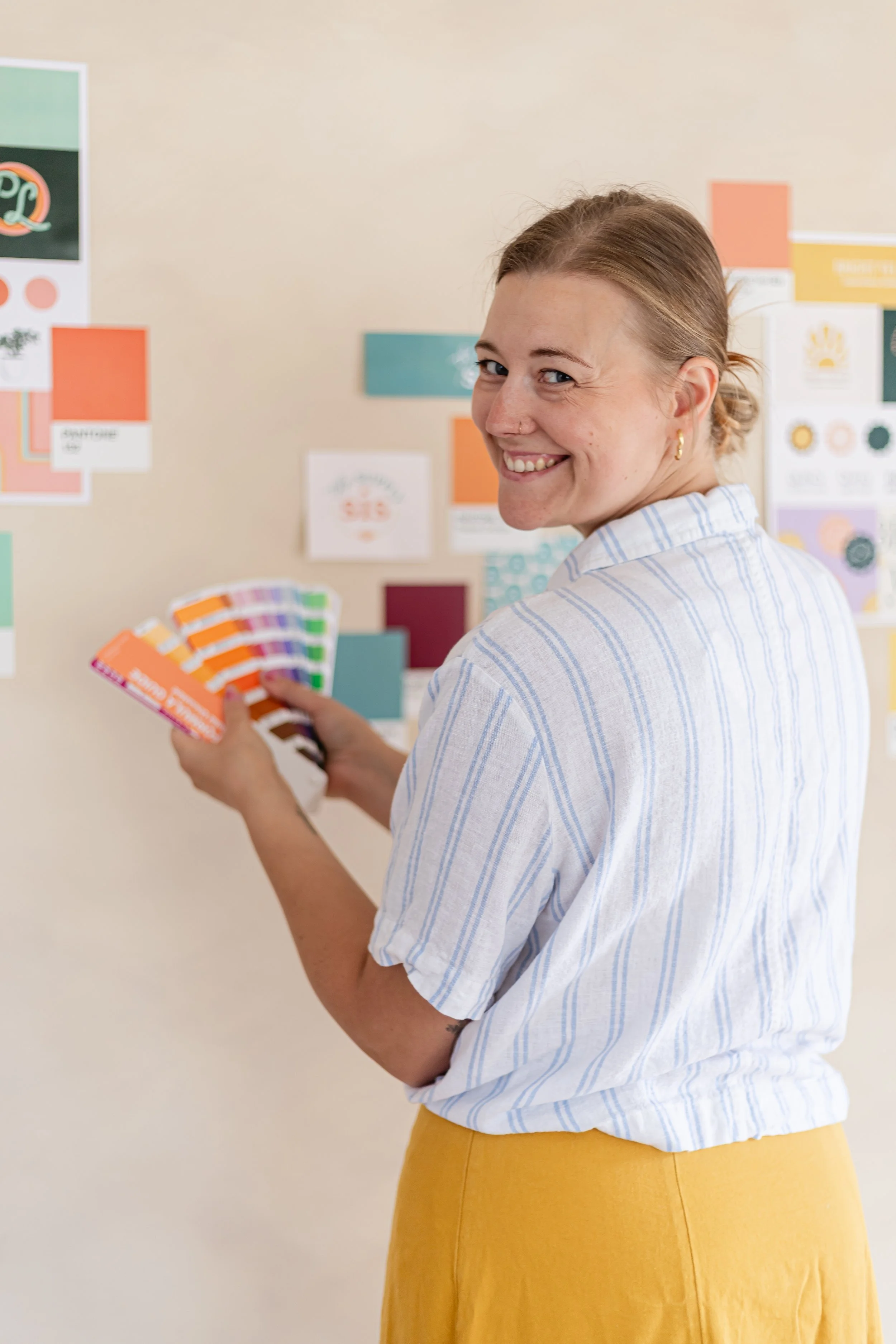 A smiling woman with blonde hair in a bun, wearing a white and blue striped shirt and yellow skirt, holding a selection of colorful paint swatches and standing in front of a wall with various color samples and design boards.