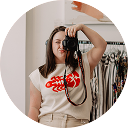 Young woman taking a mirror selfie in a clothing store fitting room, holding a camera, wearing a white t-shirt with a red graphic design, and beige pants.