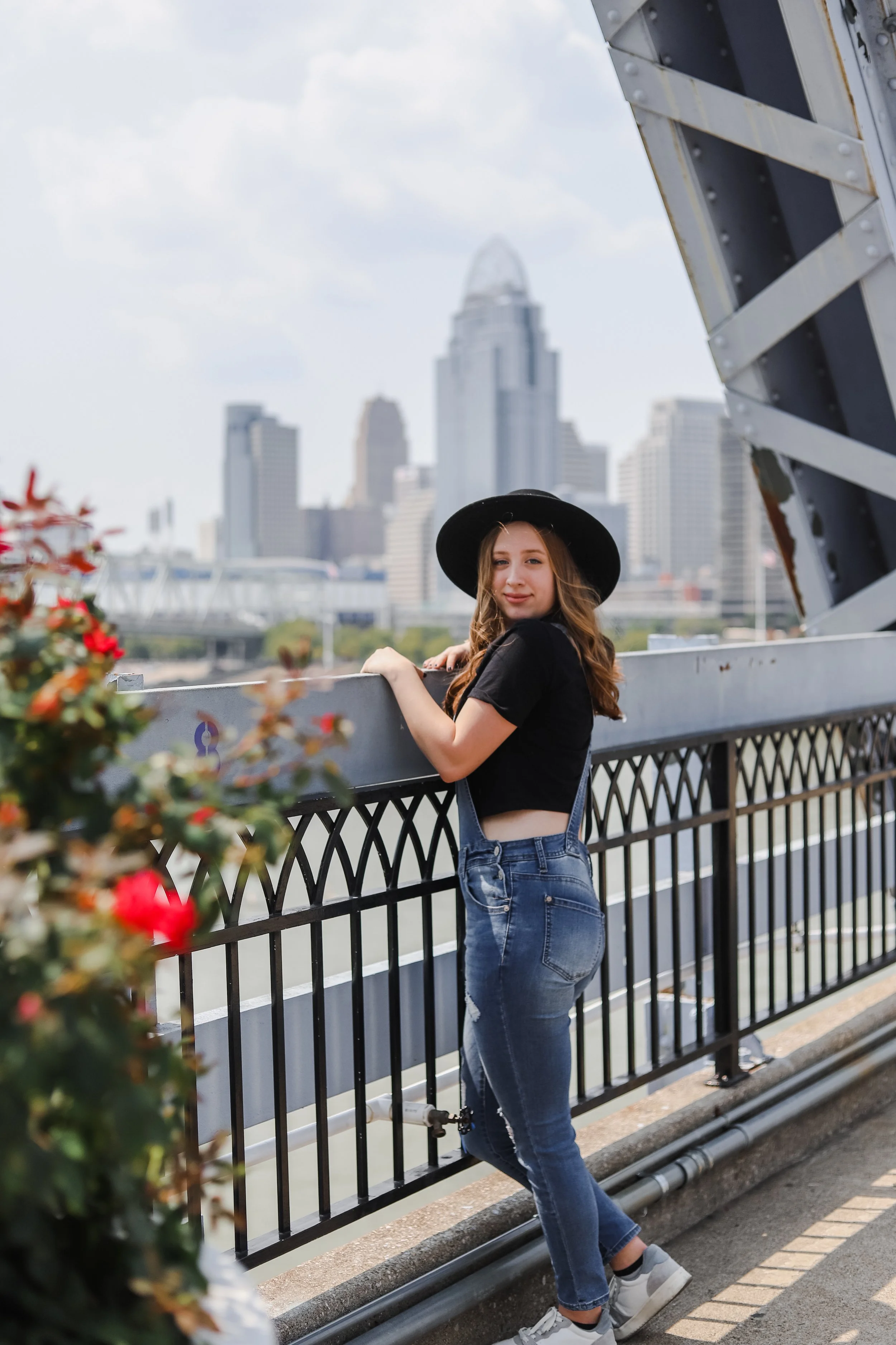 A young woman with long brown hair, wearing a black hat, black crop top, and denim overalls, stands by a metal railing with a city skyline in the background on a cloudy day.