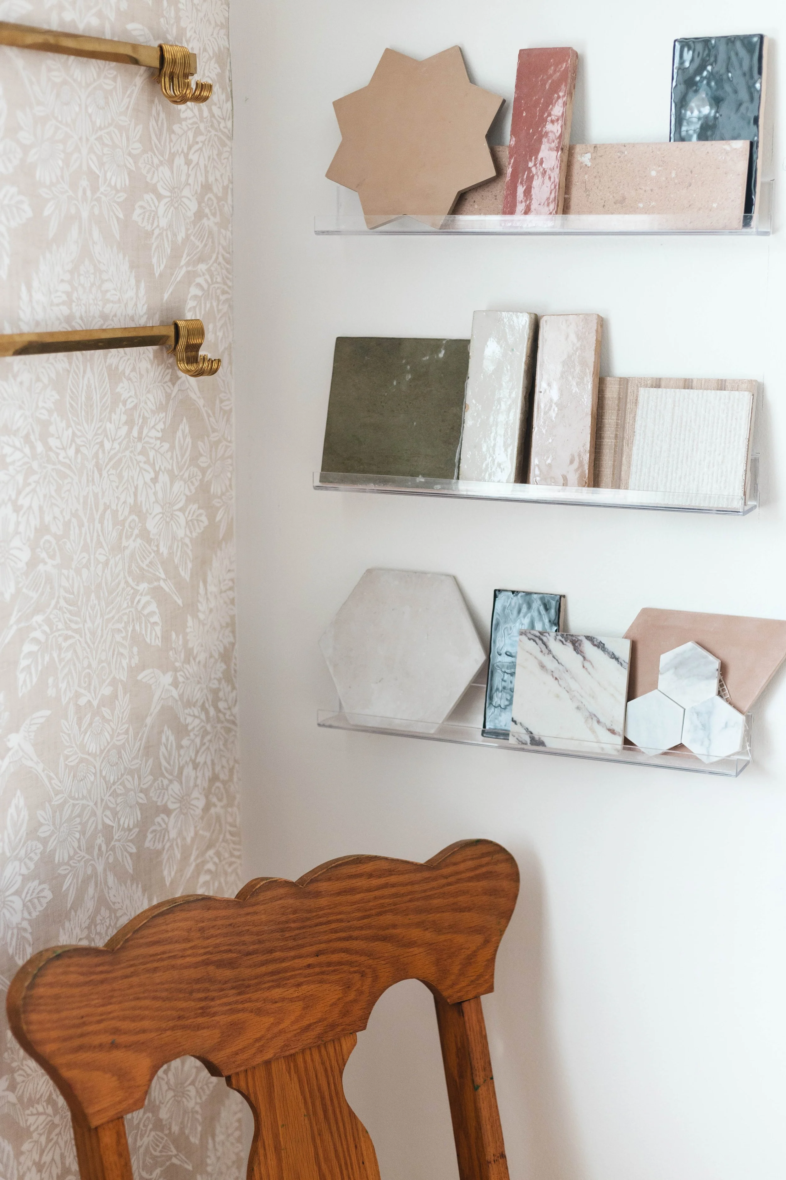 Display of various decorative tiles and stone samples on three clear wall-mounted shelves in a room with white walls and beige floral wallpaper, with part of a wooden chair visible in the foreground.