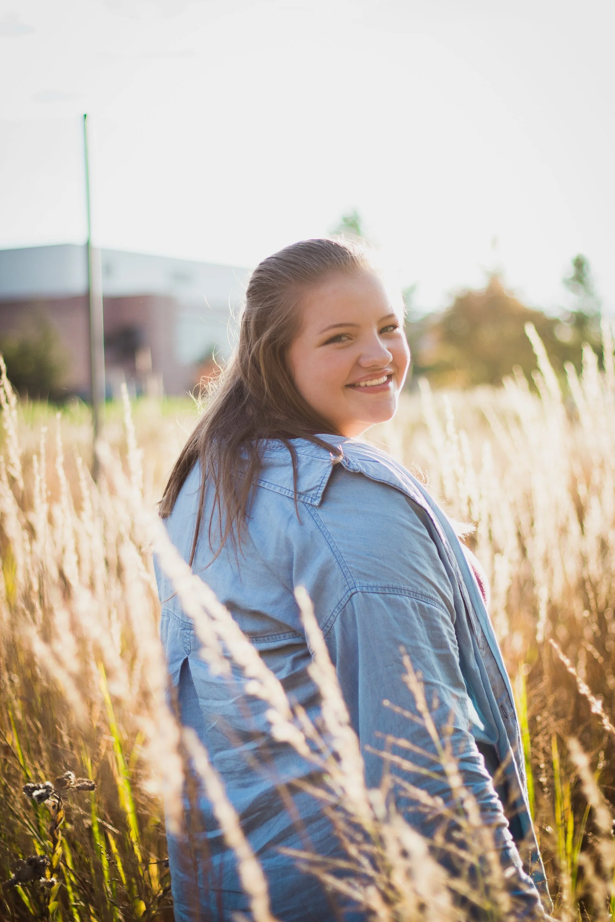 A young woman smiling, wearing a light blue jacket, standing in a field of tall grass with a building in the background during sunny weather.
