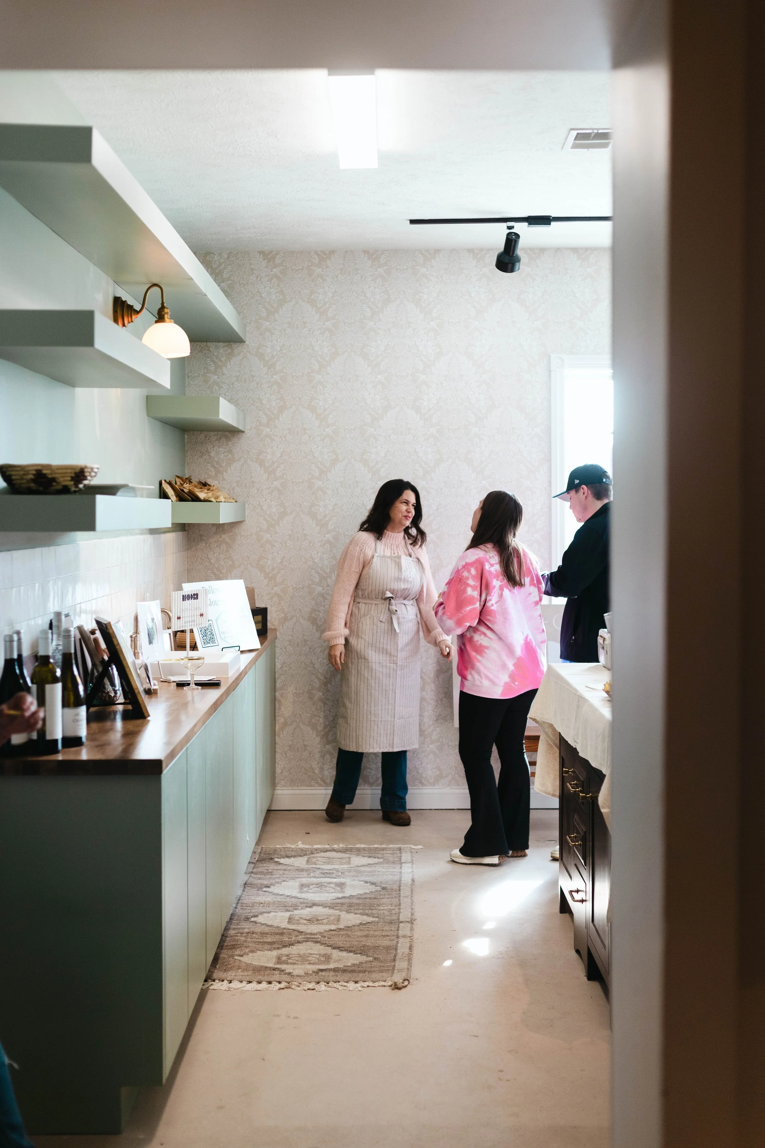 Three people standing and talking in a cozy kitchen or dining area, with kitchen counters, wine bottles, and framed photos visible on the counter.