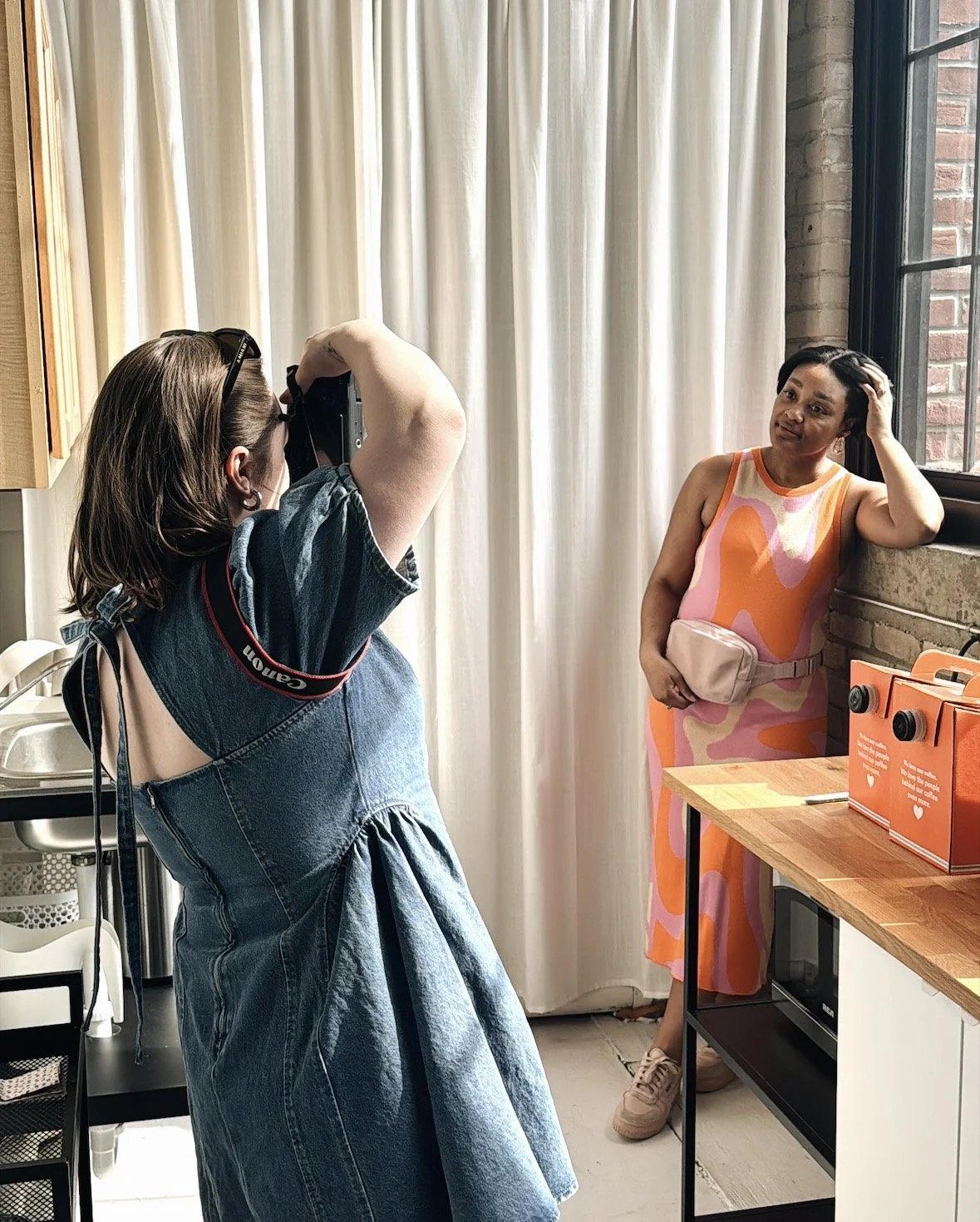 A woman taking a photo of another woman in a colorful dress inside a room with cream curtains, a brick wall, and a wooden table with orange boxes.