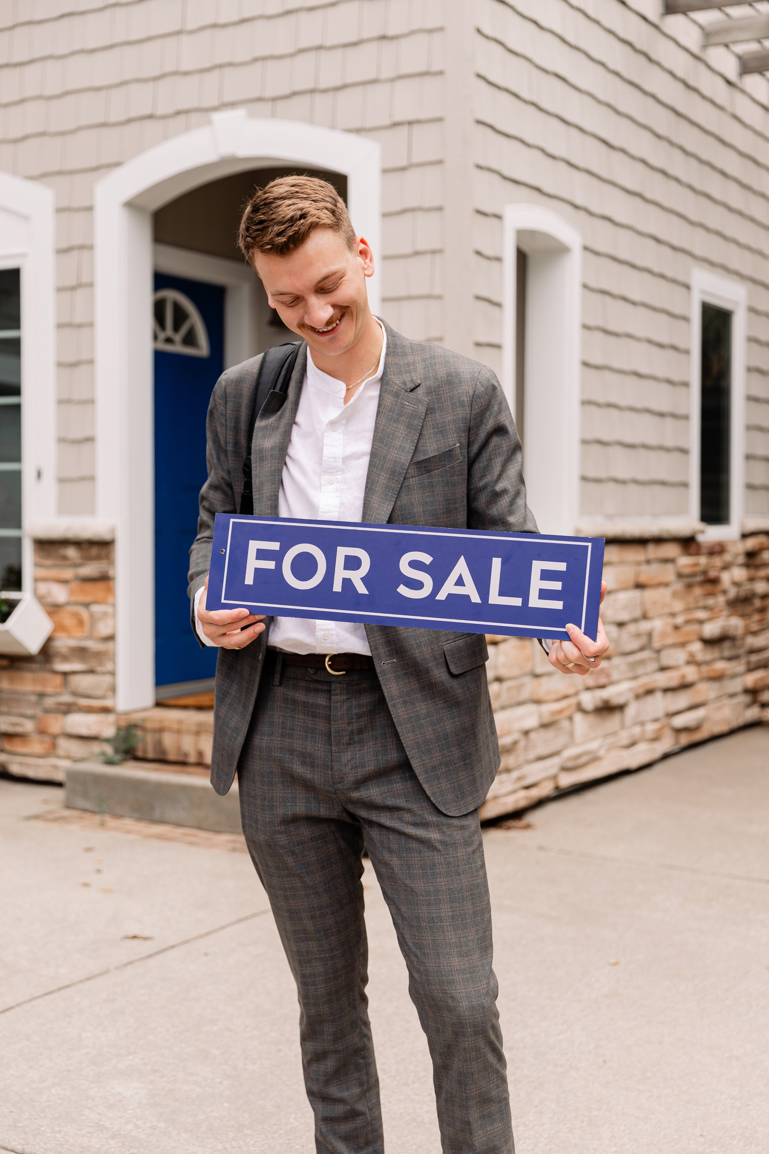 A young man in a suit holding a "For Sale" sign standing outside a house with a stone and siding exterior.