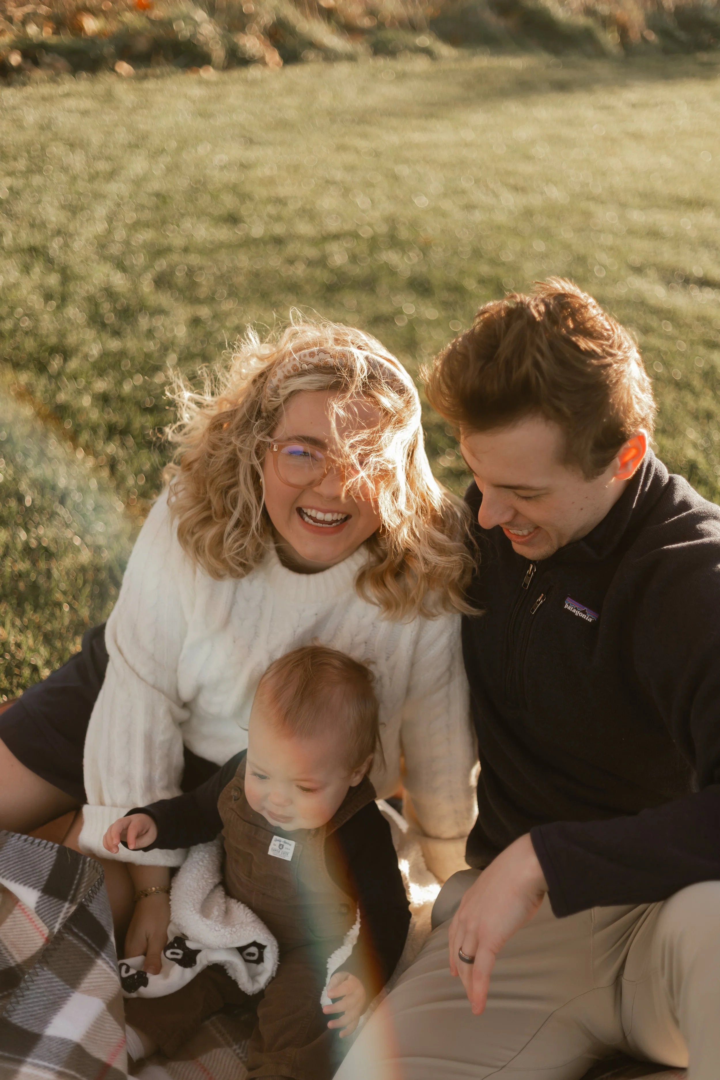 A happy family sitting on grass outdoors, with a woman, a man, and a young child, all smiling and enjoying the moment.