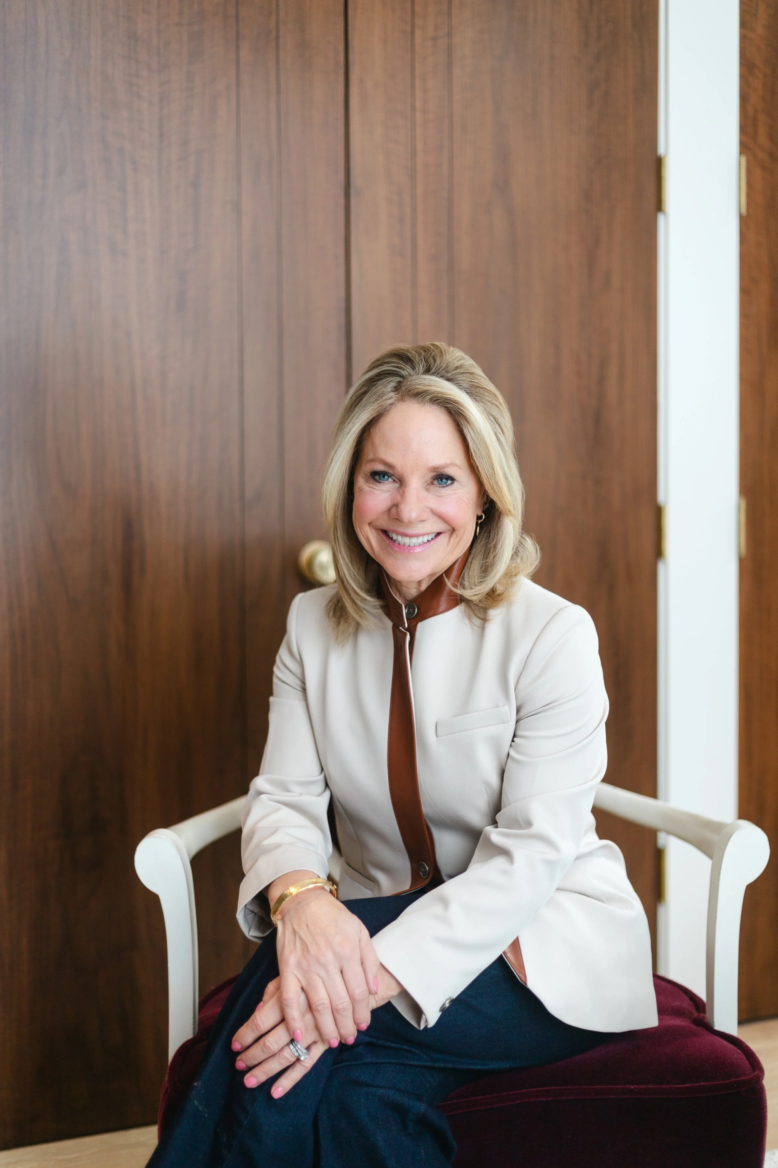 A smiling woman with blonde hair, wearing a beige blazer with brown trim, sitting on a white chair with a dark red cushion, in front of a wooden wall.
