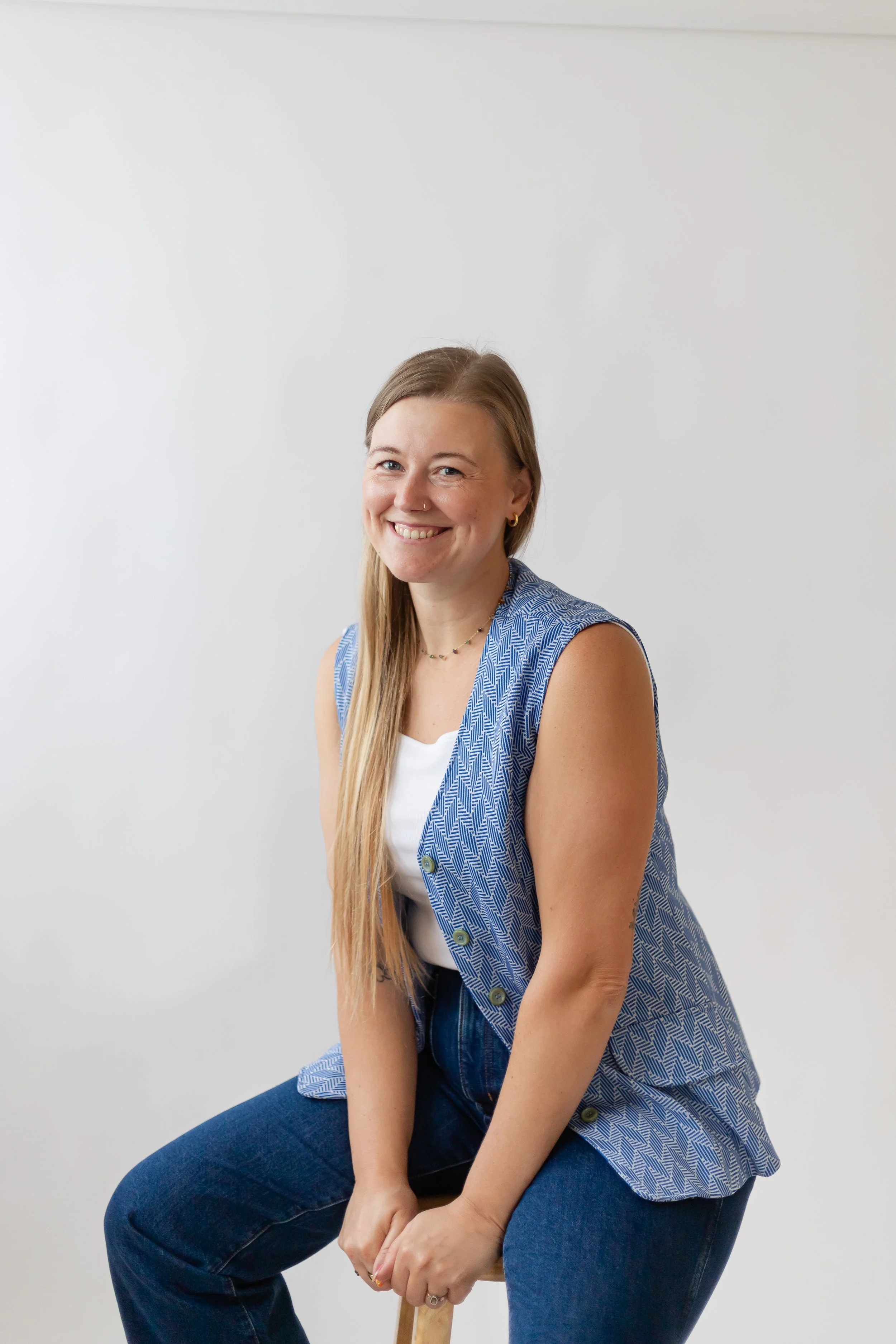 A woman with long blonde hair, smiling, sitting on a stool against a plain white background, wearing a blue patterned vest and dark jeans.