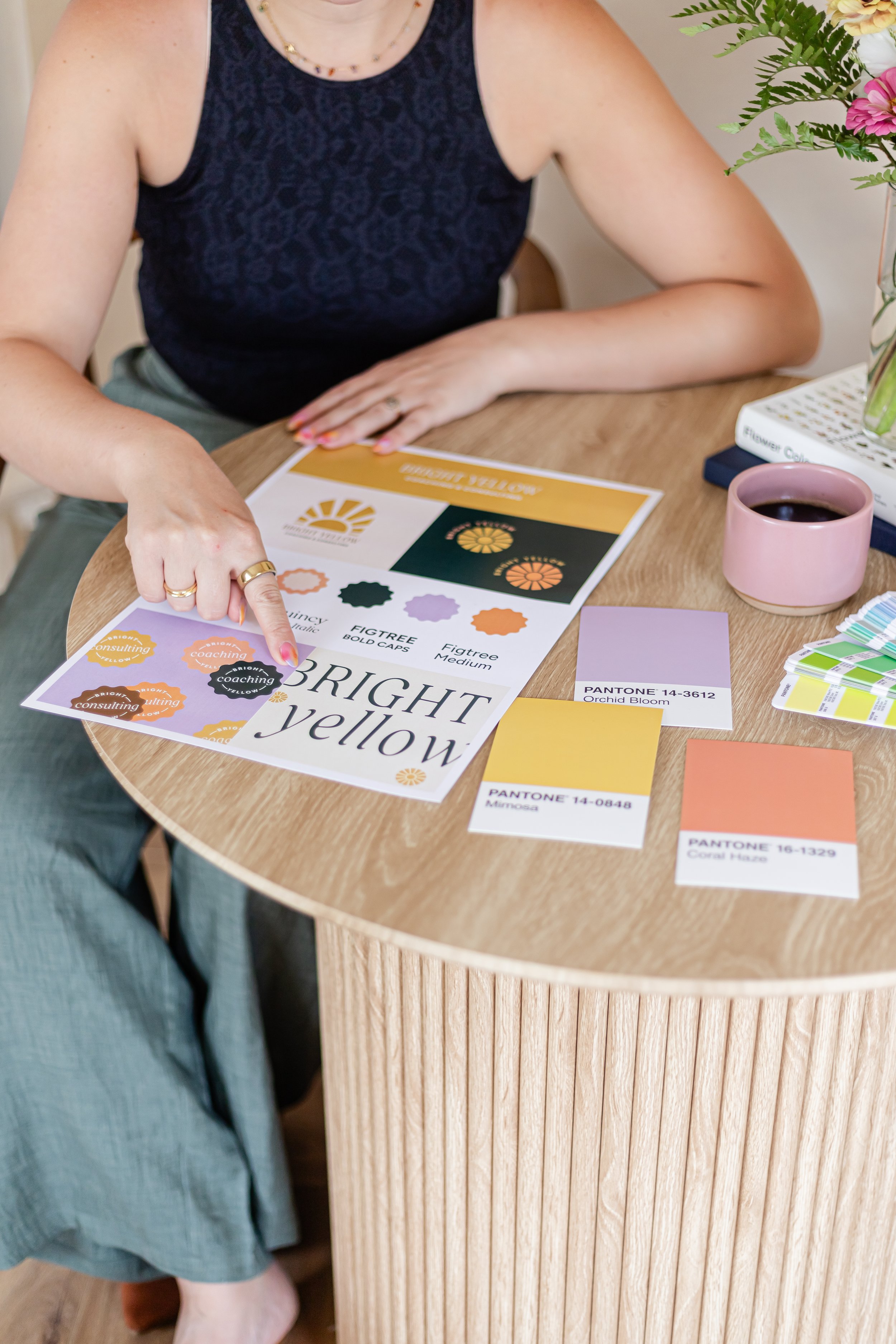 Woman sitting at a wooden table, pointing to color and design samples, with a pink cup of coffee, flower arrangement, and design magazine on the table.
