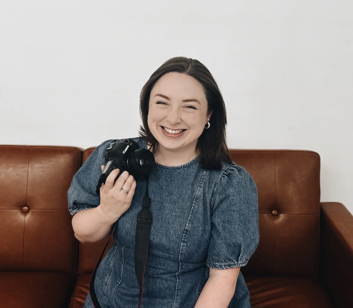 A woman smiling and holding a Canon camera while sitting on a brown leather sofa against a plain white wall.